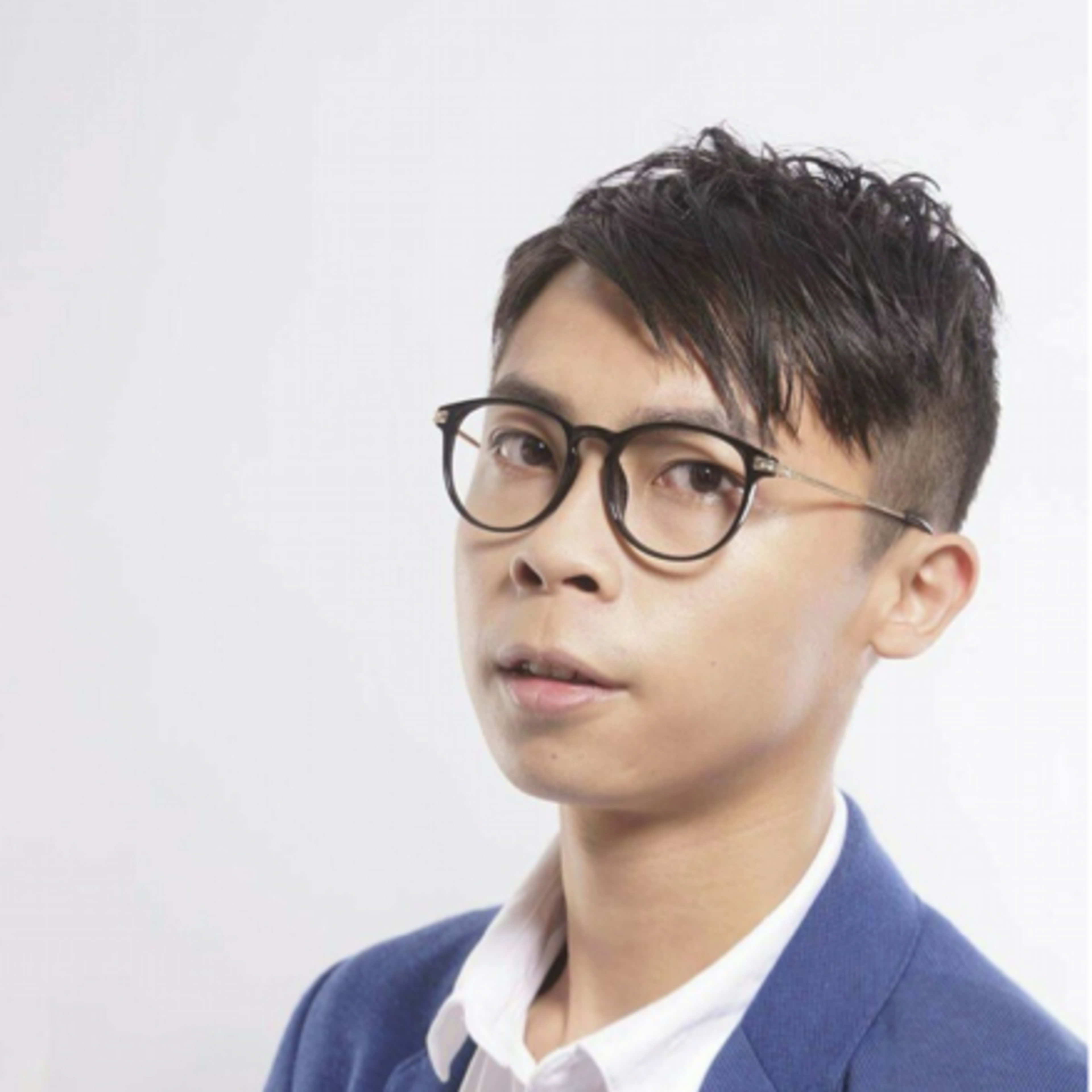 A studio headshot of Hong Kong theatre actor Chiu Chin Hei, captured against a clean, neutral background. He is wearing a sophisticated blue blazer with a white shirt and black-rimmed glasses, embodying the professional aesthetic of Rooftop Productions' contemporary stage performers. This portrait highlights his expressive presence as a key talent in the local independent theatre scene.