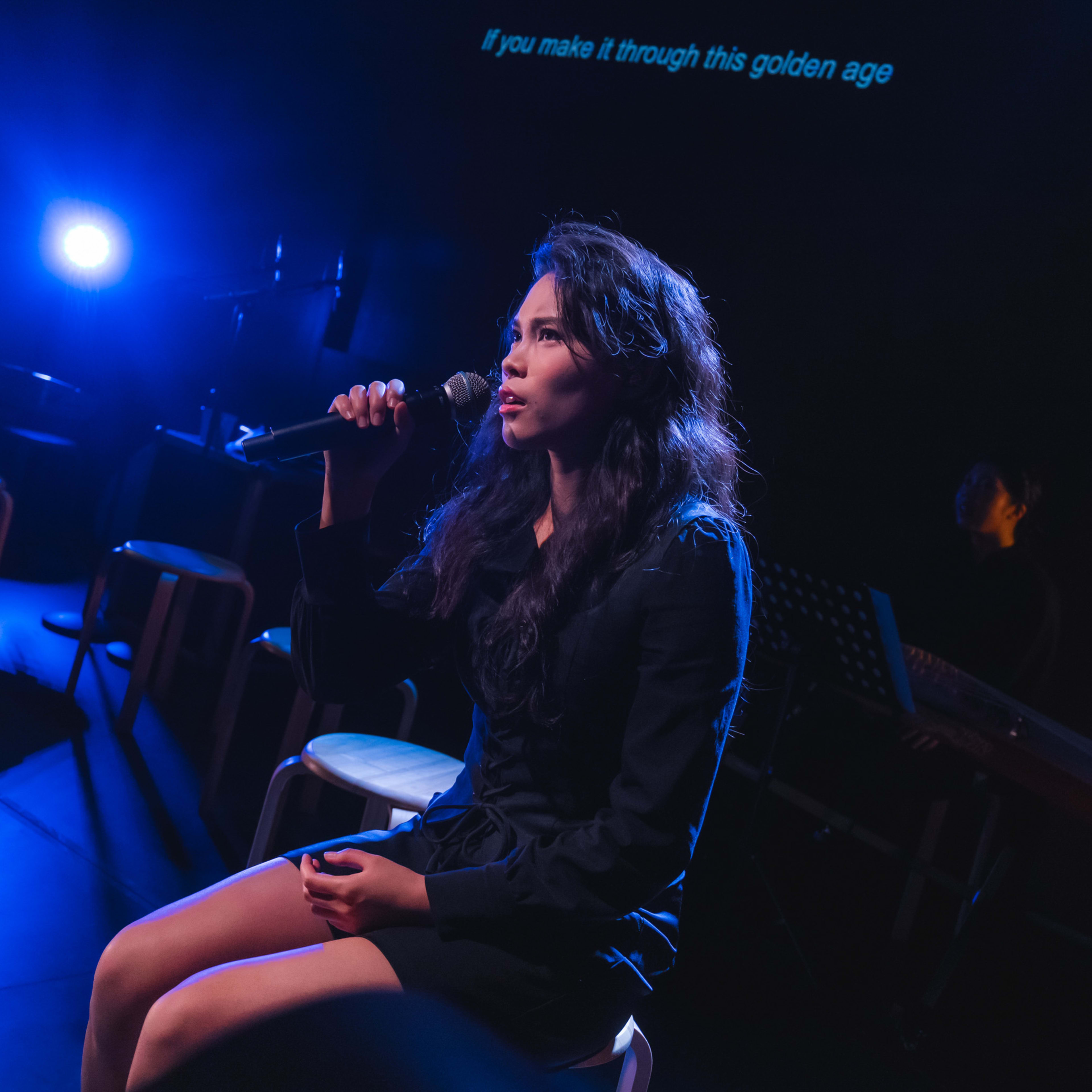 錢穎嘉在天台製作的當代香港舞台劇《曲解莎劇》中飾演 Aemilia Lanier。舞台燈光呈現戲劇性的藍色，錢穎嘉坐在高腳凳上，手持麥克風深情演唱，情感投入。舞台環境簡約且黑暗，陰影處隱約可見樂器。她唱道：「日日盡力活在大時代。」