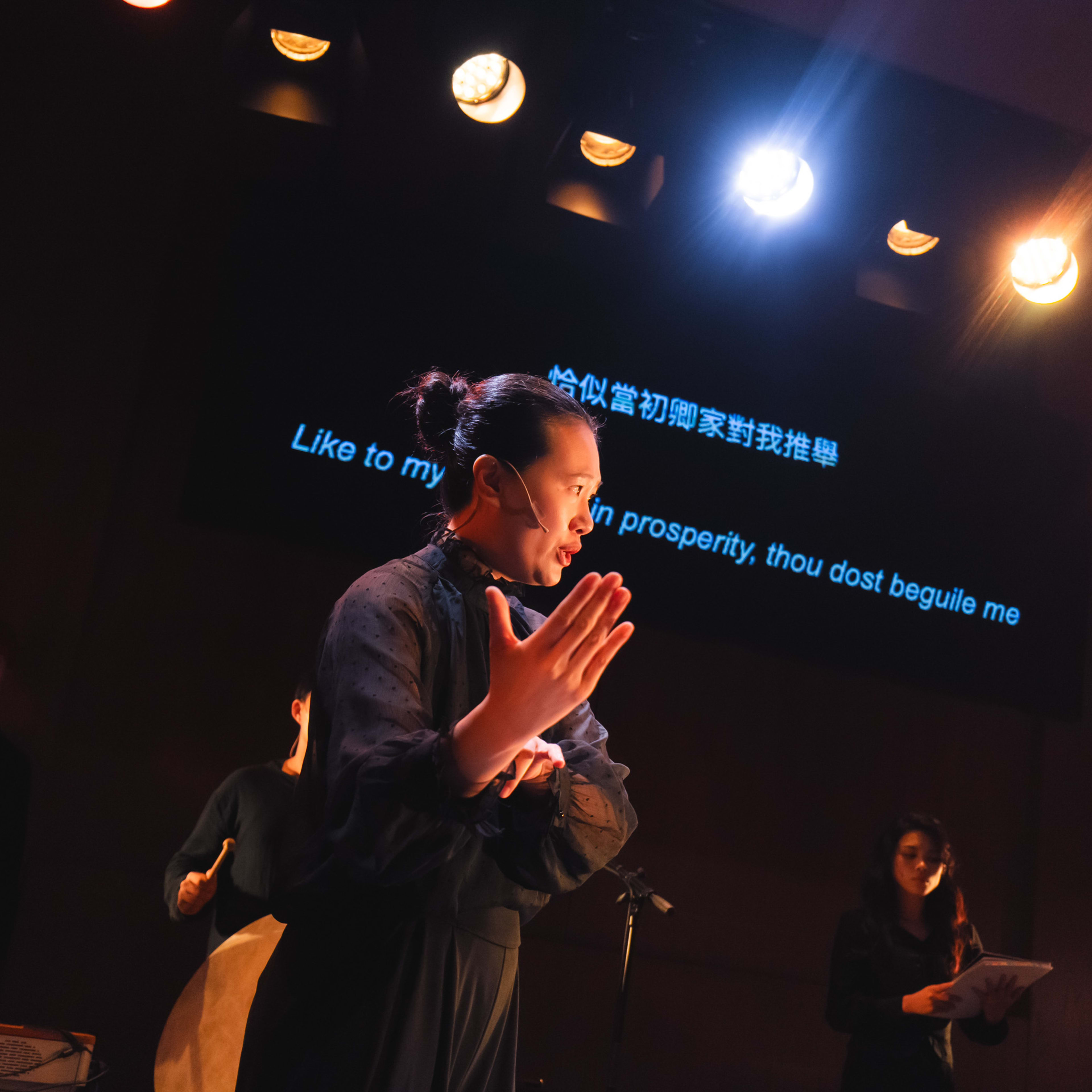 在天台製作（Rooftop Productions）的當代香港劇場作品《曲解莎劇》（History Play）中，演員錢穎嘉身處舞台中央，被暖色調的燈光籠罩。她正以埃米莉亞·蘭尼爾（Aemilia Lanier）的角色進行演出，手勢充滿張力，身後的螢幕投影出劇中對白「恰似當初卿家對我推舉」。現場的戲劇燈光營造出深沉的情感氛圍。錢穎嘉飾演的埃米莉亞·蘭尼爾念道：「恰似當初卿家對我推舉」