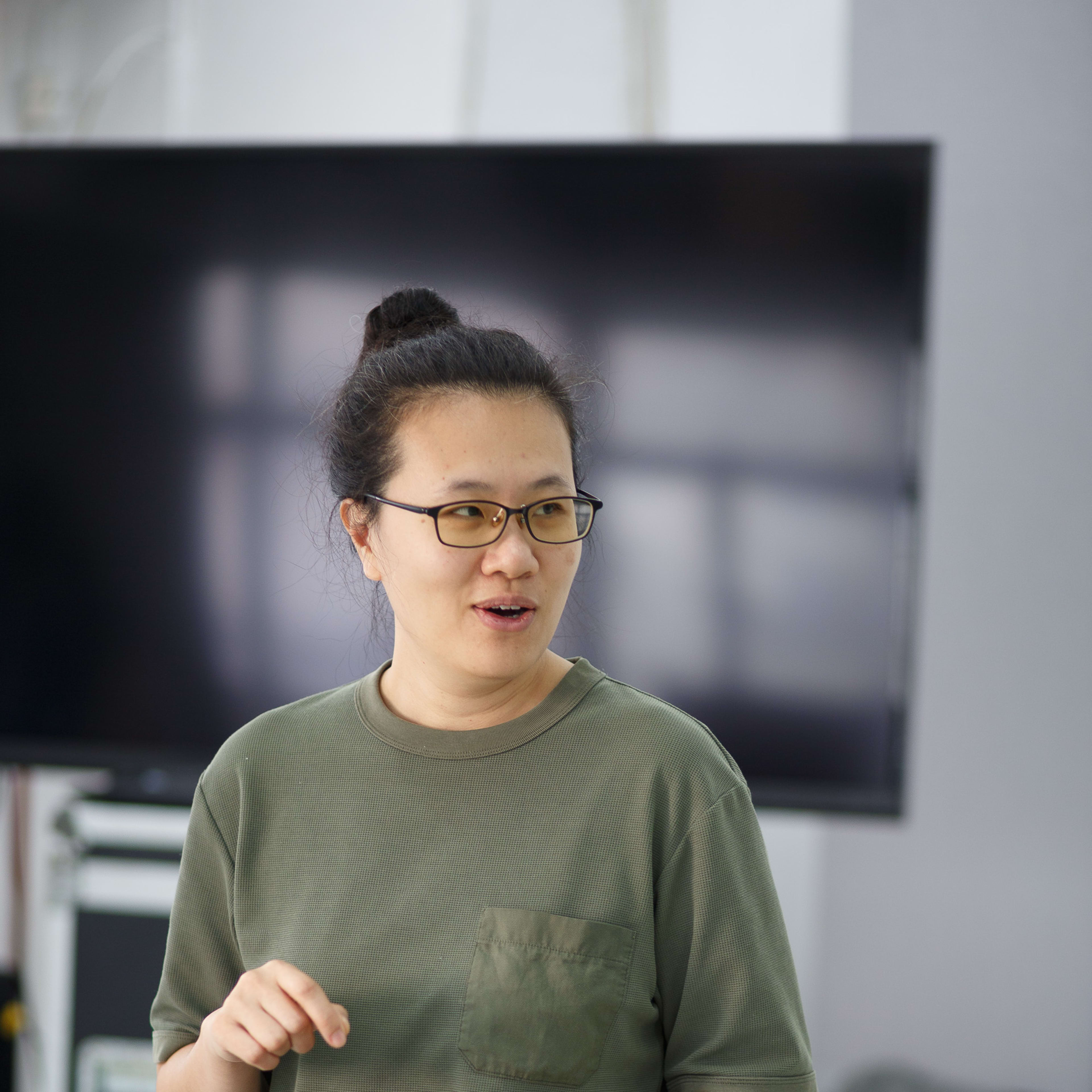 演員李婉晶於香港當代劇場作品《卡啦OK》的排練現場中專注演繹。她身穿休閒橄欖綠色T恤並戴著眼鏡，雙手配合表演動作，表情生動，背景為排練室及屏幕。此為《卡啦OK》排練現場。
