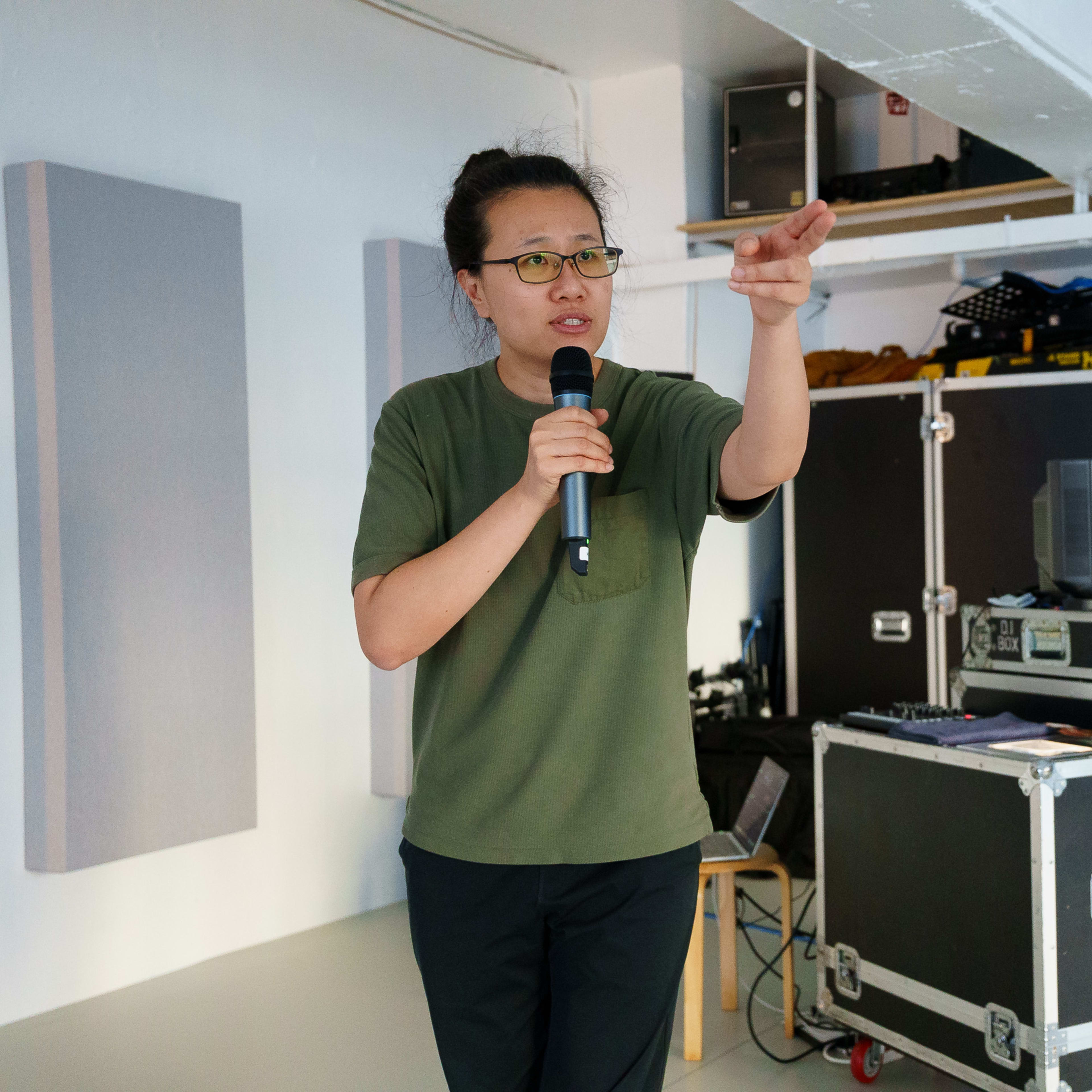 演員李婉晶在天台製作的當代劇場作品《卡啦OK》排練現場，手持麥克風並指向前方，身後擺放著演出用的飛行箱和器材。《卡啦OK》排練。