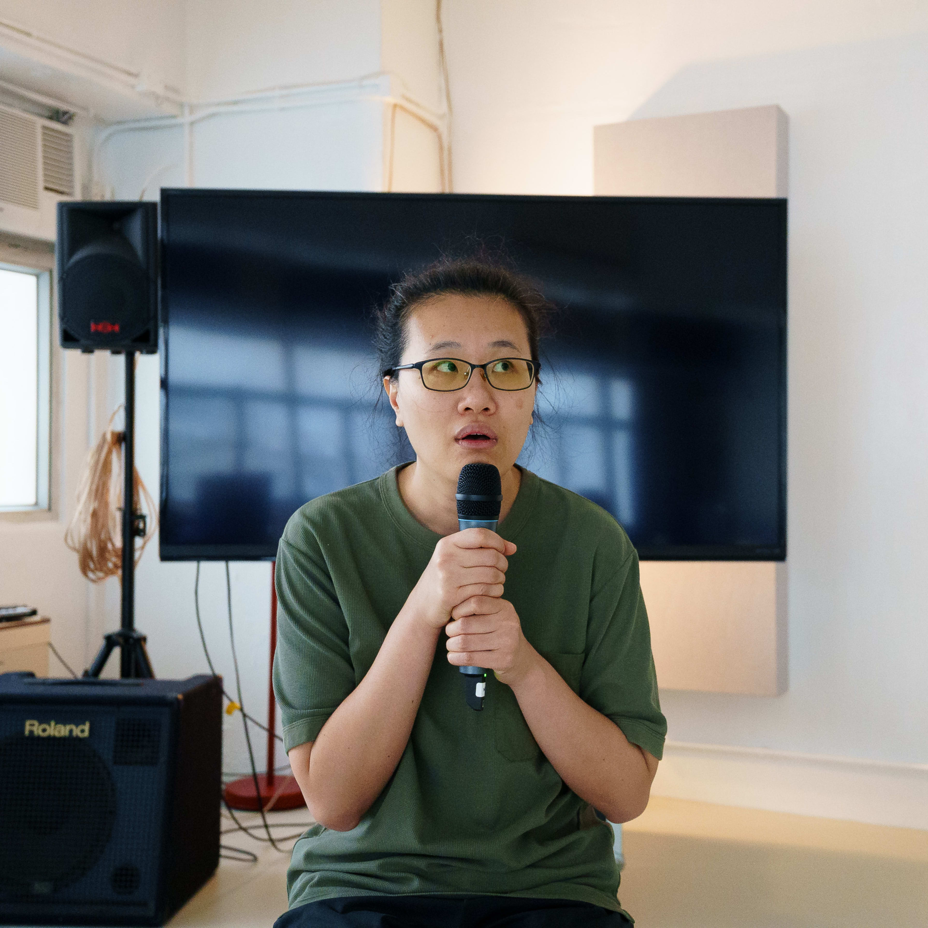 李婉晶於天台製作（Rooftop Productions）的當代劇場作品《卡啦OK》排練期間手持麥克風。她身處設備完善的排練室中，背景可見Roland擴音器與音響設備，呈現出表演者專注投入的創作狀態。《卡啦OK》排練