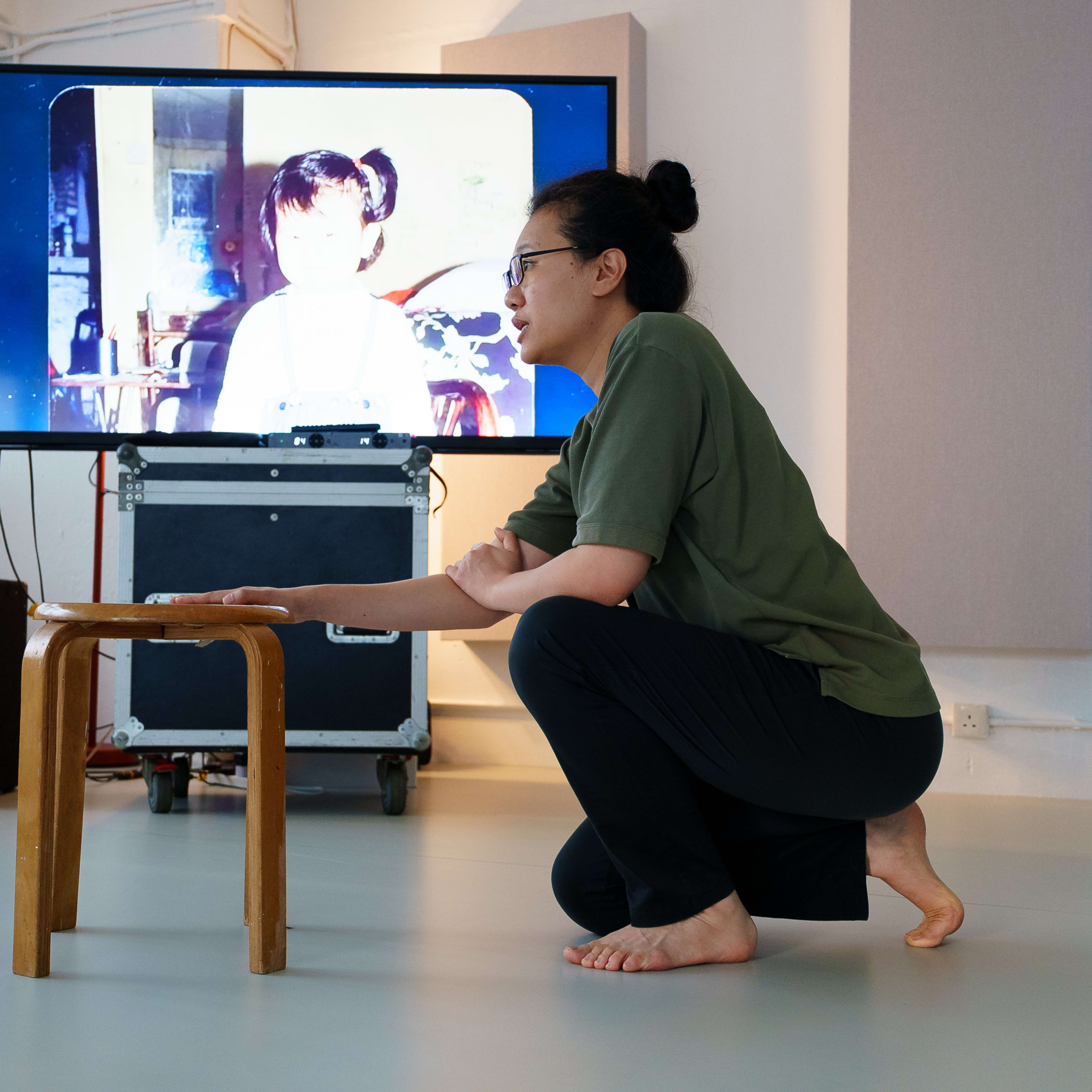 在天台製作 (Rooftop Productions) 當代劇場作品《卡啦OK》的排練現場，演員李婉晶正沉浸於角色之中。她身著舒適的排練服，單膝跪地，手扶著木凳，神情專注而細膩。身後的螢幕映照著一張充滿懷舊感的幼年女孩圖像，營造出探討記憶與表演的劇場氛圍。攝影記錄下了這次專業的舞台排練過程。《卡啦OK》排練。
