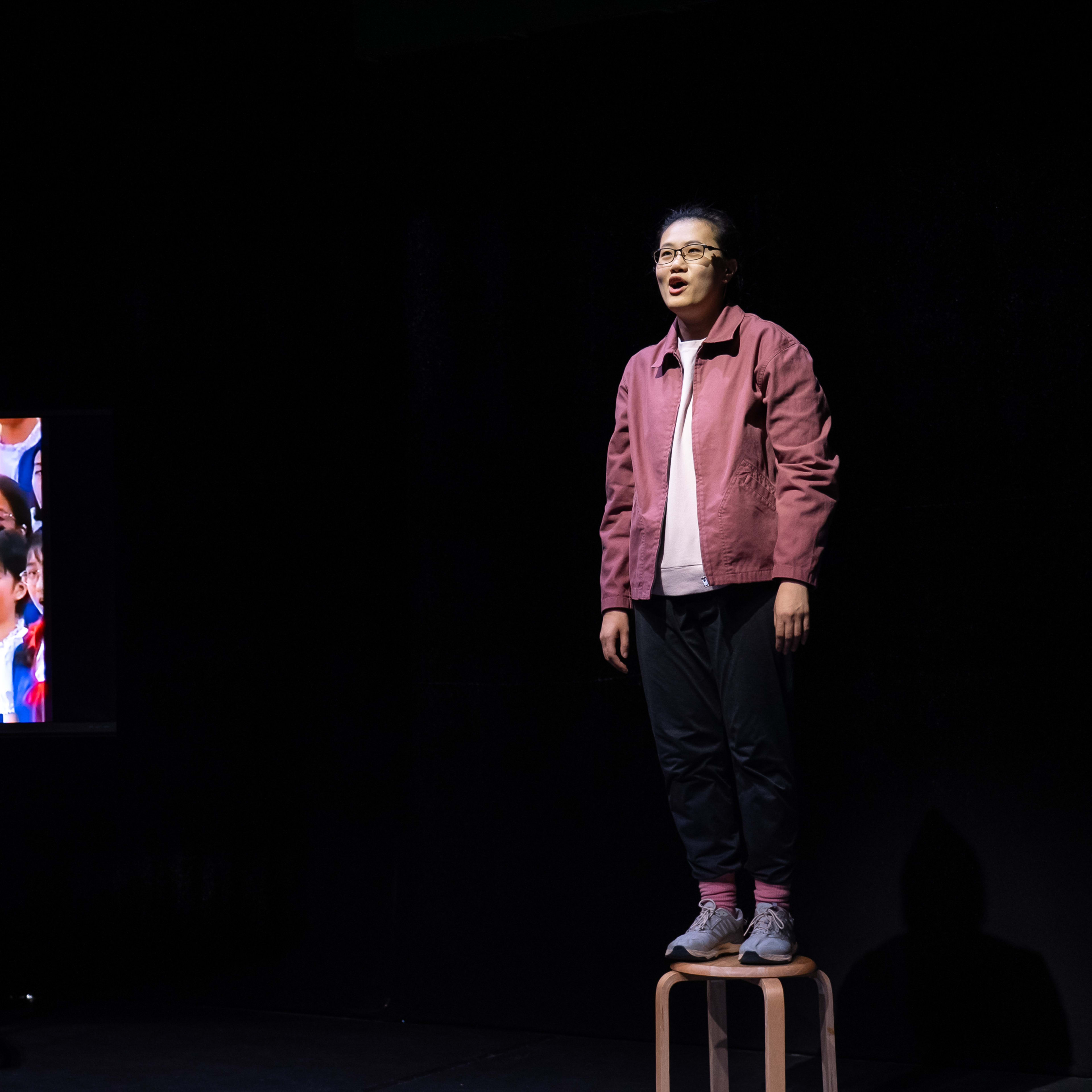 在天台製作的當代劇場作品《卡啦OK》的極簡舞台上，演員李婉晶站在一張小凳子上。她身穿休閒的紫紅色外套並戴著眼鏡，表情投入地進行演出，眼神略帶仰望。舞台左側的顯示屏播放著一張張年輕臉孔的合照，營造出充滿懷舊感的檔案影像。這一幕展現了該劇對記憶與身份議題的深刻探討。李婉晶唱道：「世上所有的兒童。」