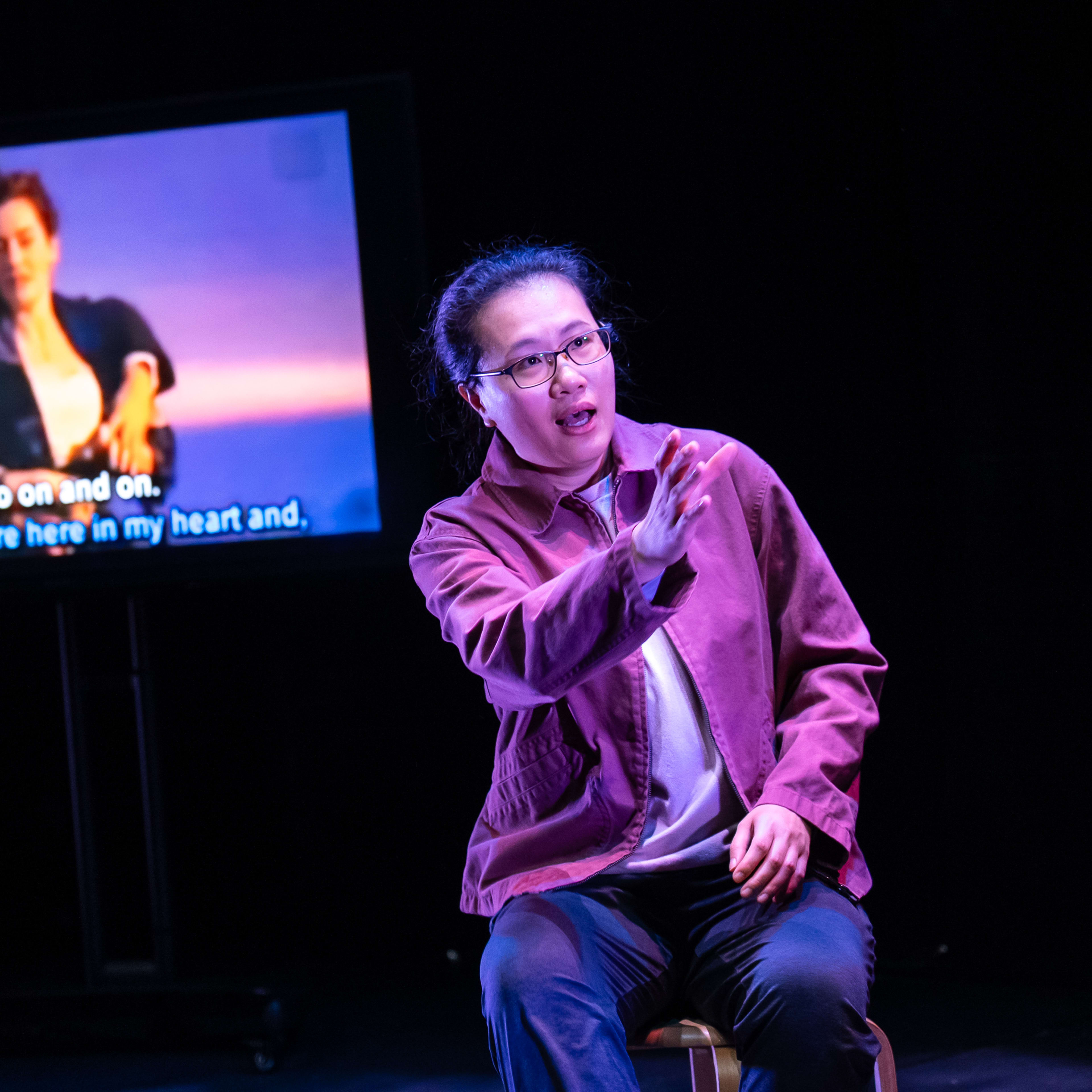 在天台製作的當代香港劇場作品《卡啦OK》中，演員李婉晶坐在舞台中央的凳子上，身處強烈的舞台燈光與全黑背景之間。她伸出手臂，表情流露出深沉而細膩的情感。身後的螢幕正播放電影《鐵達尼號》的經典浪漫劇照，畫面下方的歌詞字幕與舞台氛圍相互映襯。她深情演繹道：「不管是遠是近，無論你在哪裏。」