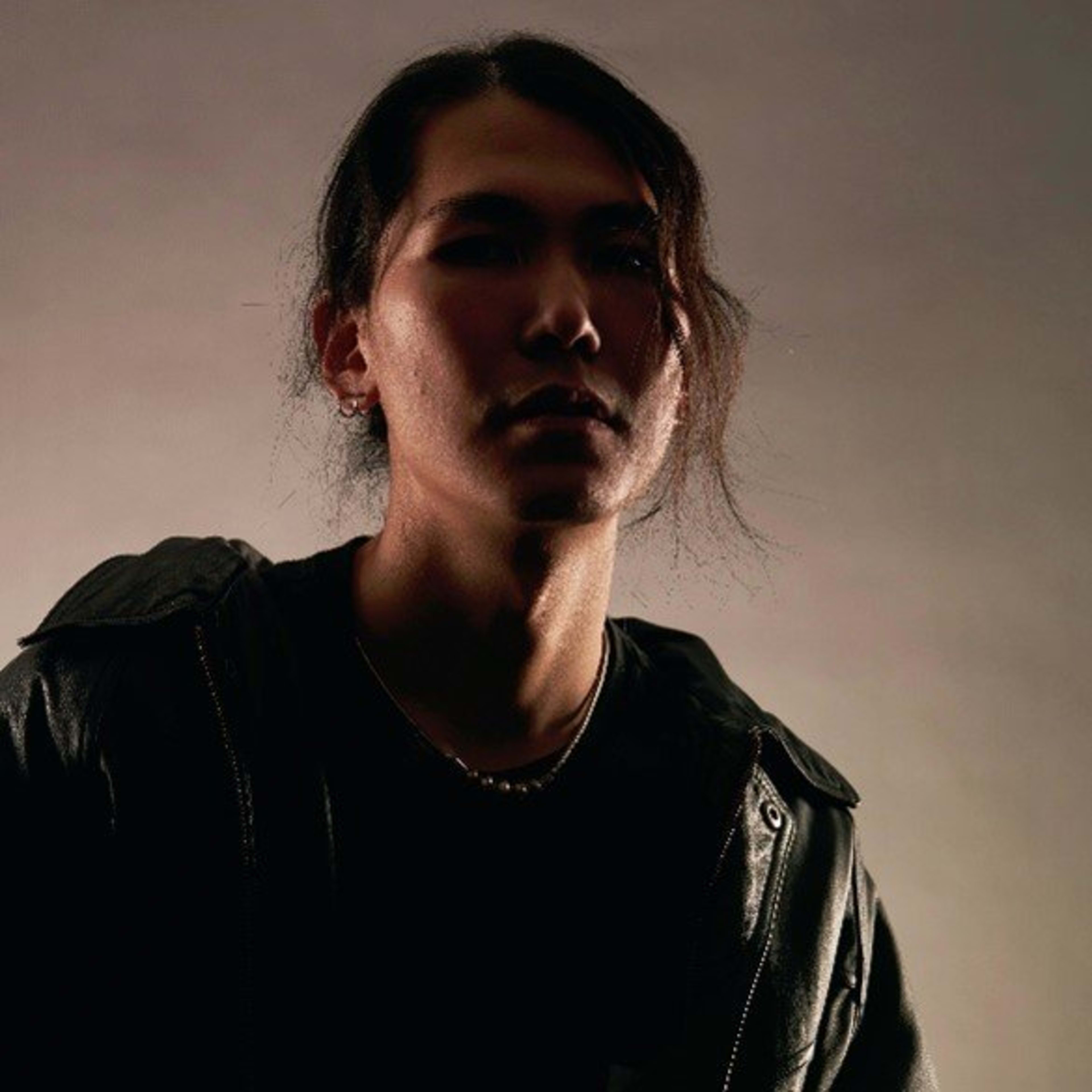 A dramatic studio headshot of Hong Kong actor Lau Hiu Kong (Lawrence) for Rooftop Productions (天台製作). He is captured in a medium close-up, wearing a dark leather jacket against a neutral backdrop. The lighting creates high-contrast shadows that highlight his intense, contemplative expression and textured hair, embodying the moody, contemporary aesthetic of modern HK theatre performance art.