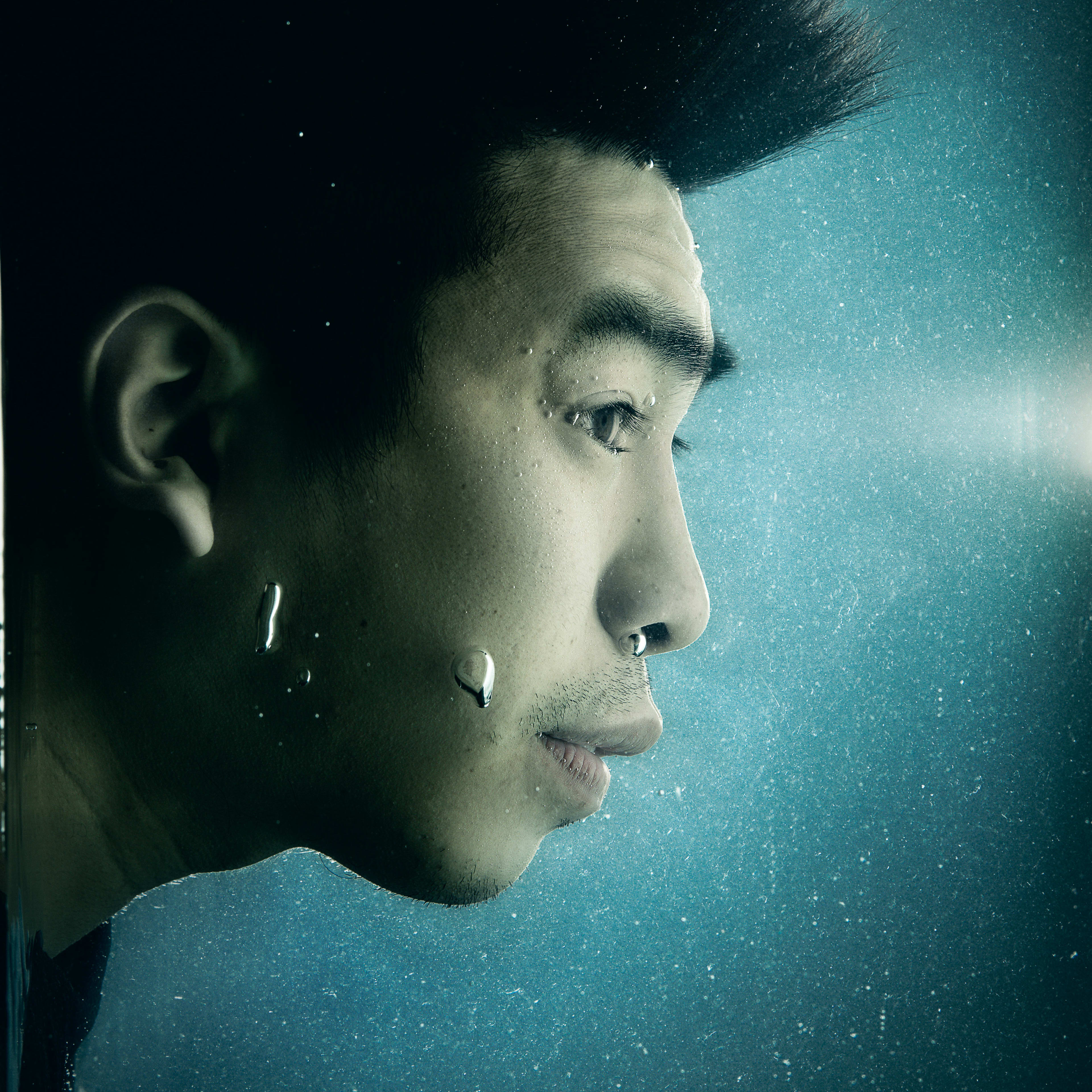 A dramatic close-up profile shot of actor Larry Ng submerged underwater, his hair floating upward in a spiky silhouette against a dark, moody blue background. Small air bubbles cling to his skin and drift through the water, creating a contemplative atmosphere typical of Rooftop Productions' avant-garde stage aesthetic. This promotional image for the contemporary Hong Kong theatre production 'Lost Shoreline' captures the tension and introspection of the piece. 'Lost Shoreline'