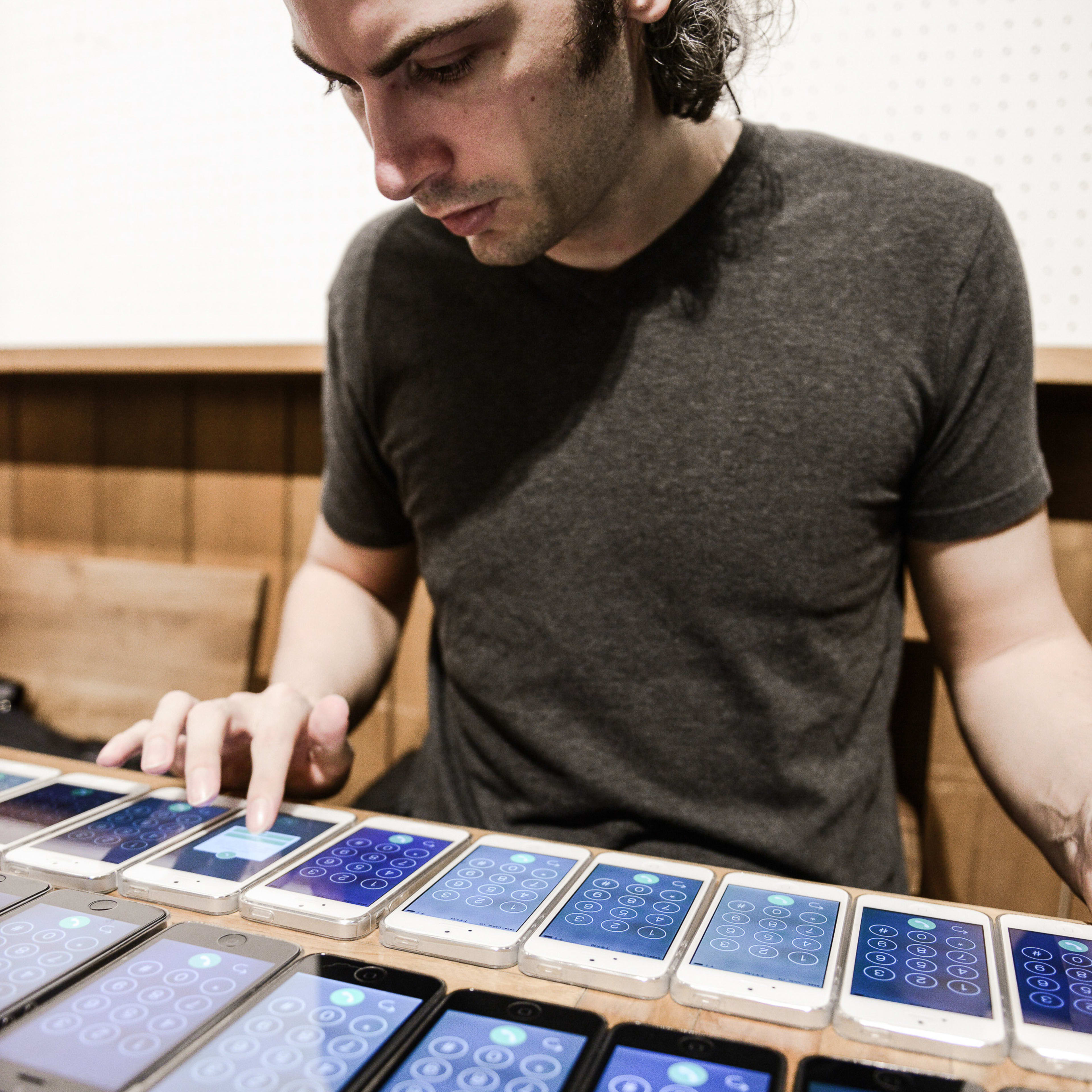 In a rehearsal for the contemporary Hong Kong theatre production Lost Shoreline, actor Ivor Houlker is captured in a focused, intimate moment. He sits at a wooden table, his hand delicately touching one of the many smartphones arranged in a precise grid before him. The screens display a digital interface, reflecting a fusion of technology and performance art. The lighting is soft and subdued, emphasizing the performer's concentration as he navigates the interactive elements of this experimental work. 'Lost Shoreline'