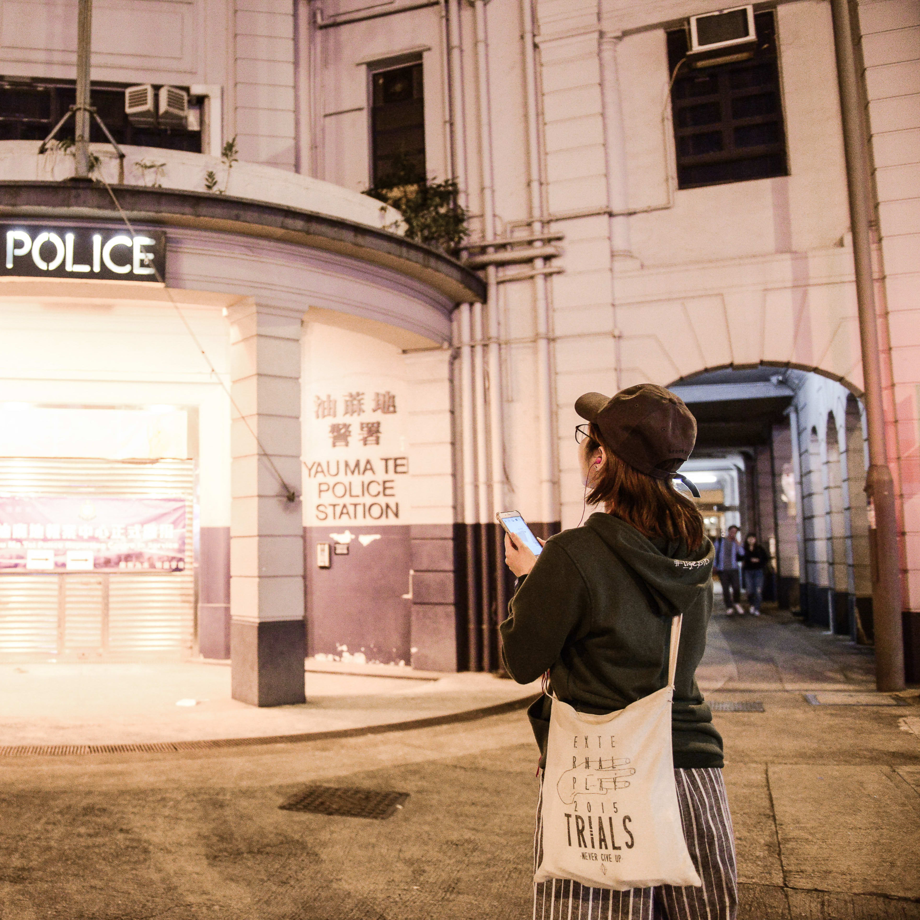 A night shot showing a person wearing a cap and hoodie standing on a street in Hong Kong, holding a phone while facing the historic, illuminated entrance of the Yau Ma Tei Police Station. This evocative scene sets the tone for Rooftop Productions' contemporary theatre piece, Lost Shoreline, which explores the shifting landscapes and history of the city. Lost Shoreline.