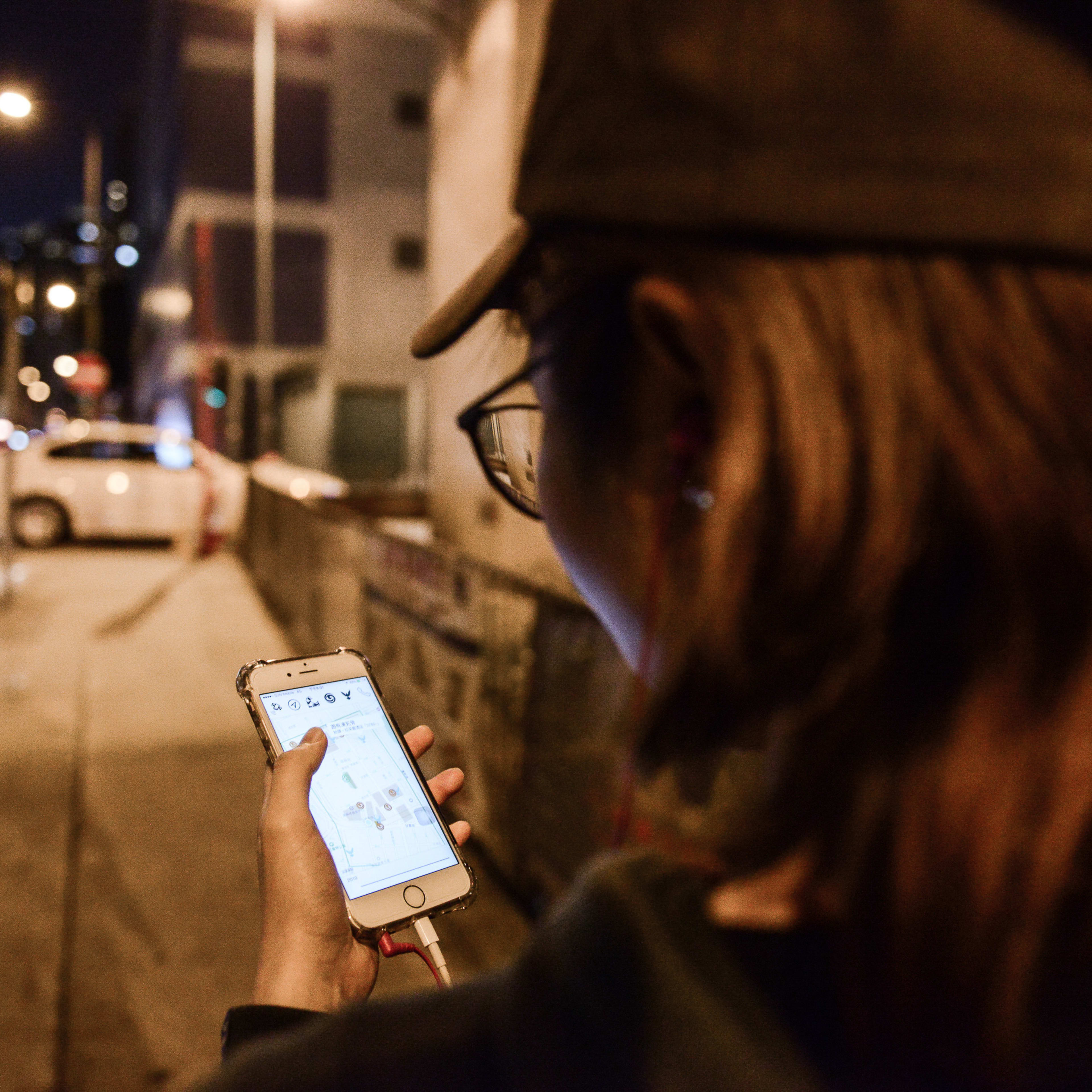 Over the shoulder shot of a person wearing a cap and glasses standing on a dark Hong Kong street at night, holding a smartphone that illuminates their hand and the surrounding area. The screen displays a digital map or document, highlighting themes of urban exploration and navigation often explored in contemporary HK theatre productions like Rooftop Productions' 'Lost Shoreline' (消失的海岸線). 'Lost Shoreline'
