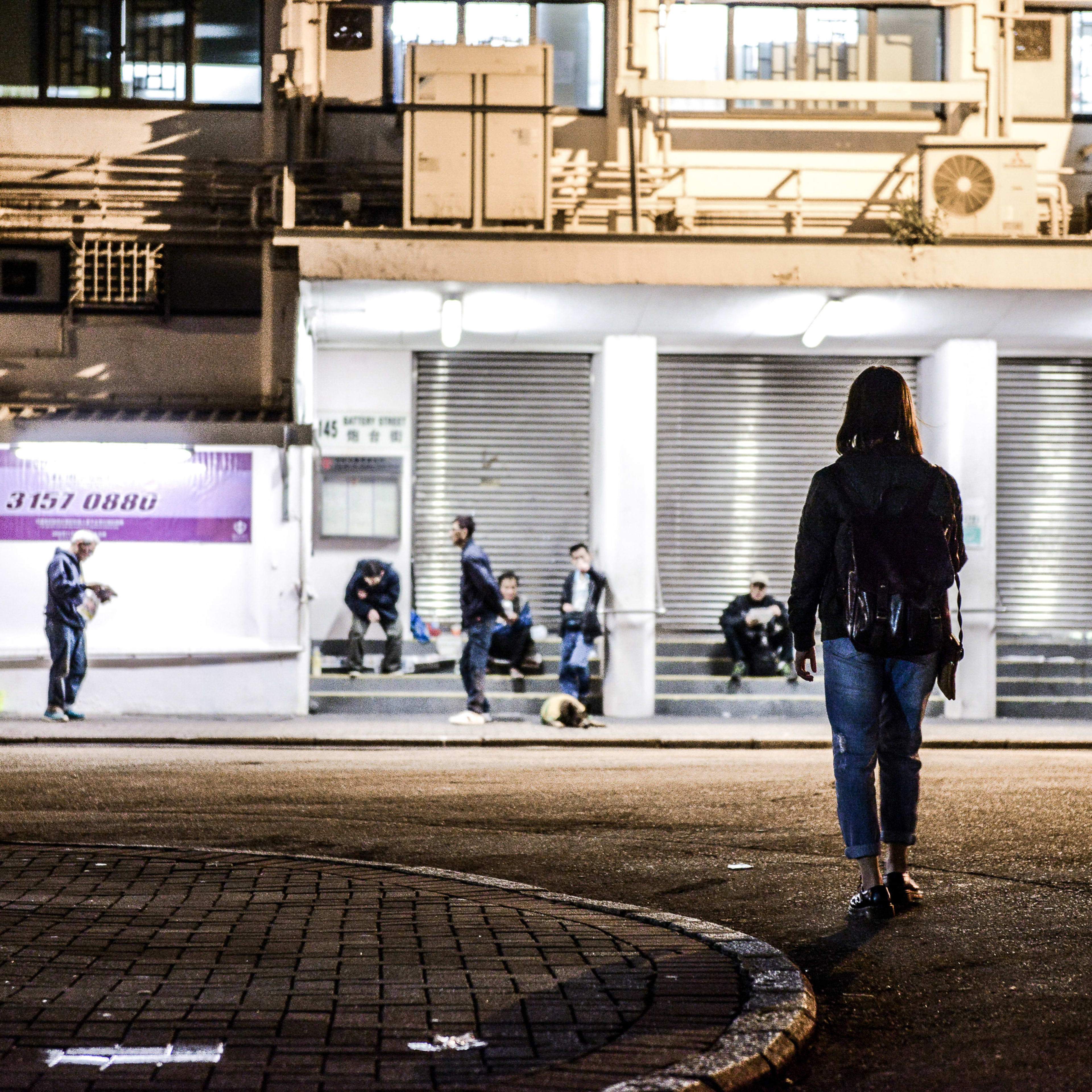 A night scene captured in a Hong Kong urban setting, featuring a person in the foreground walking away from the camera toward a brightly lit building entrance where several figures sit on the steps. The industrial architectural backdrop and the quiet, solitary atmosphere reflect the haunting, contemplative themes of Rooftop Productions' contemporary stage play, Lost Shoreline (《消失的海岸線》). Lost Shoreline