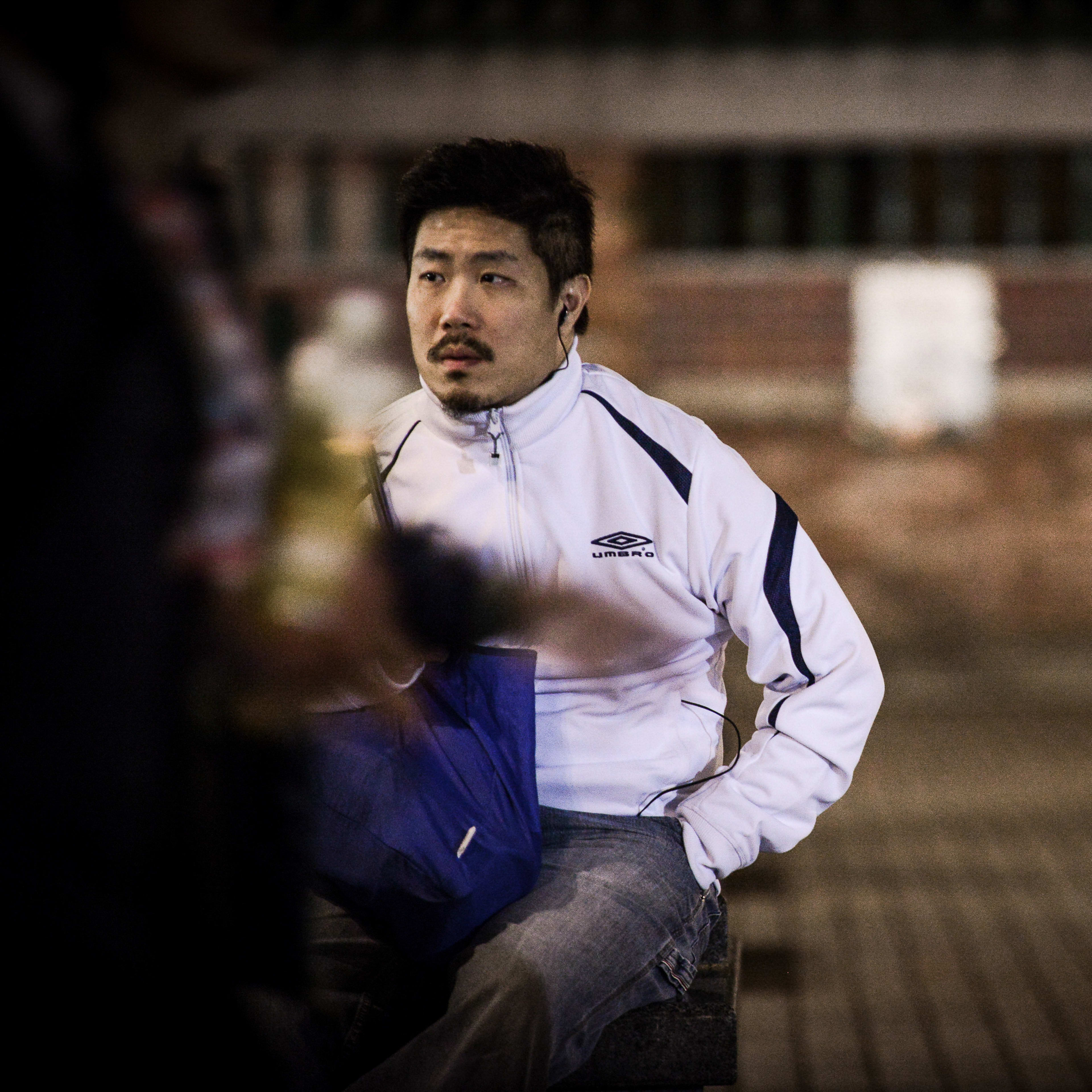 In this rehearsal capture for the Rooftop Productions (天台製作) contemporary theatre piece 'Lost Shoreline' (《消失的海岸線》), actor Sung Boon Ho (宋本浩) sits in a dim, urban outdoor setting. Clad in a white Umbro athletic jacket, he looks pensively to his left with a neutral, contemplative expression. The shallow depth of field softly blurs the background, where another person is visible, emphasizing the solitary and introspective atmosphere characteristic of this Hong Kong stage production. 'Lost Shoreline'