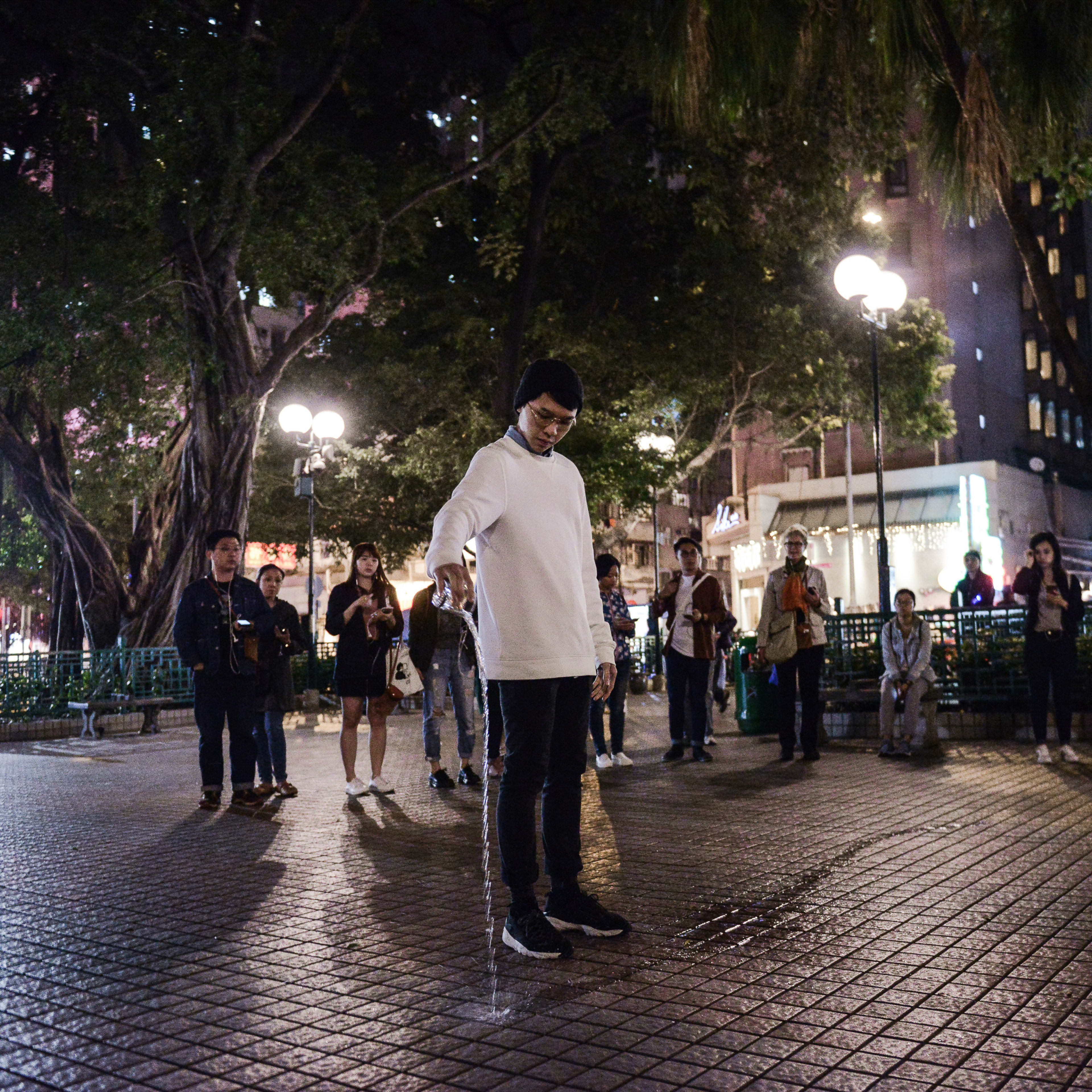 In a dimly lit outdoor park in Hong Kong, performer Ng King Lung stands centered in a white long-sleeved shirt, carefully pouring water from a small container onto the tiled ground, a poignant performance moment from the contemporary site-specific theatre production Lost Shoreline. He is surrounded by a group of observers in a semi-circle, with lush trees and urban city lights framing the background, highlighting the themes of memory and vanishing landscapes typical of Rooftop Productions' immersive work. "Lost Shoreline"
