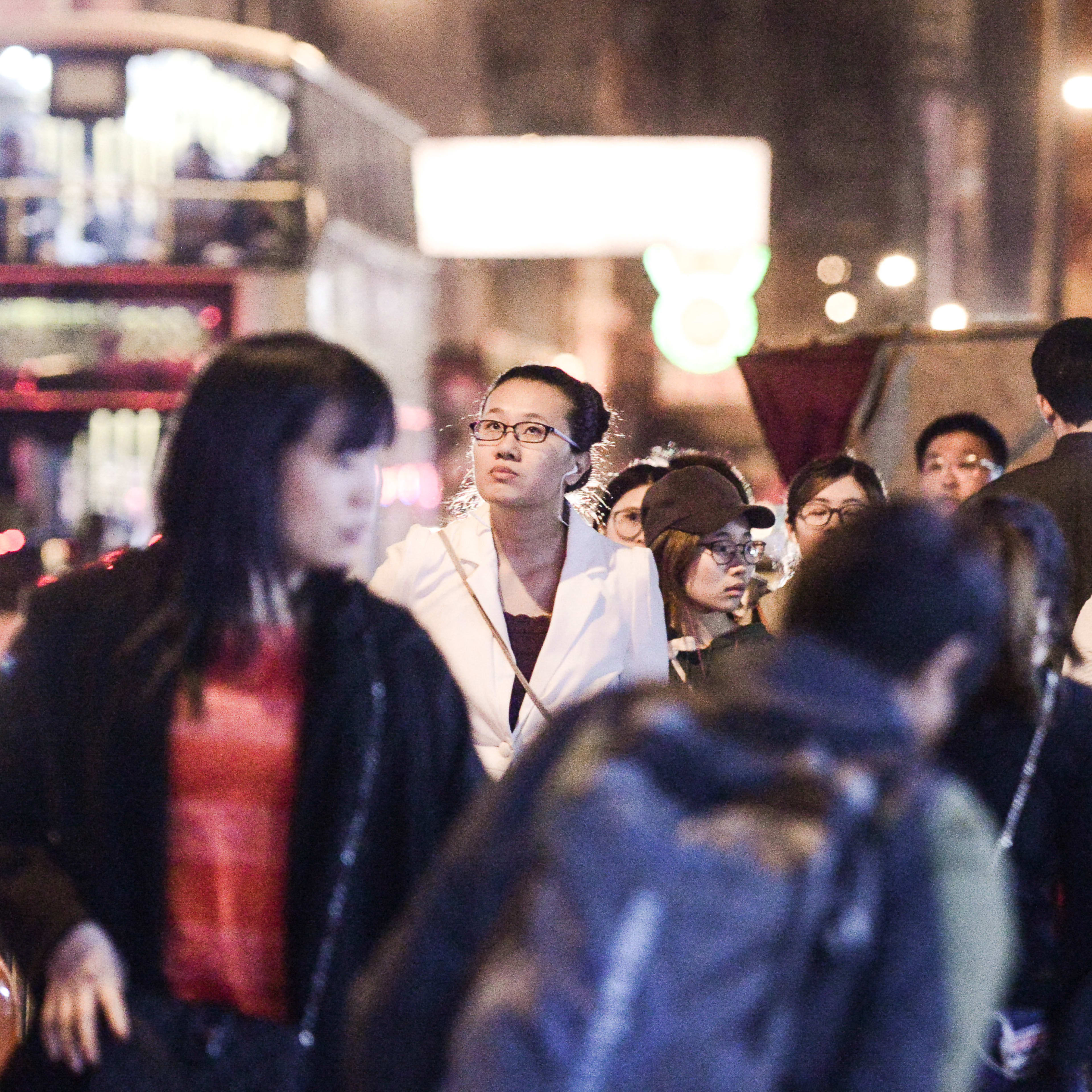 A candid, atmospheric street scene captured at night, featuring Hong Kong theatre performer Michelle Li as she gazes thoughtfully into the distance amidst a bustling crowd. This scene from Rooftop Productions’ (天台製作) contemporary theatre piece, 'Lost Shoreline', highlights the urban isolation and sensory experience of Hong Kong life. 'Lost Shoreline'