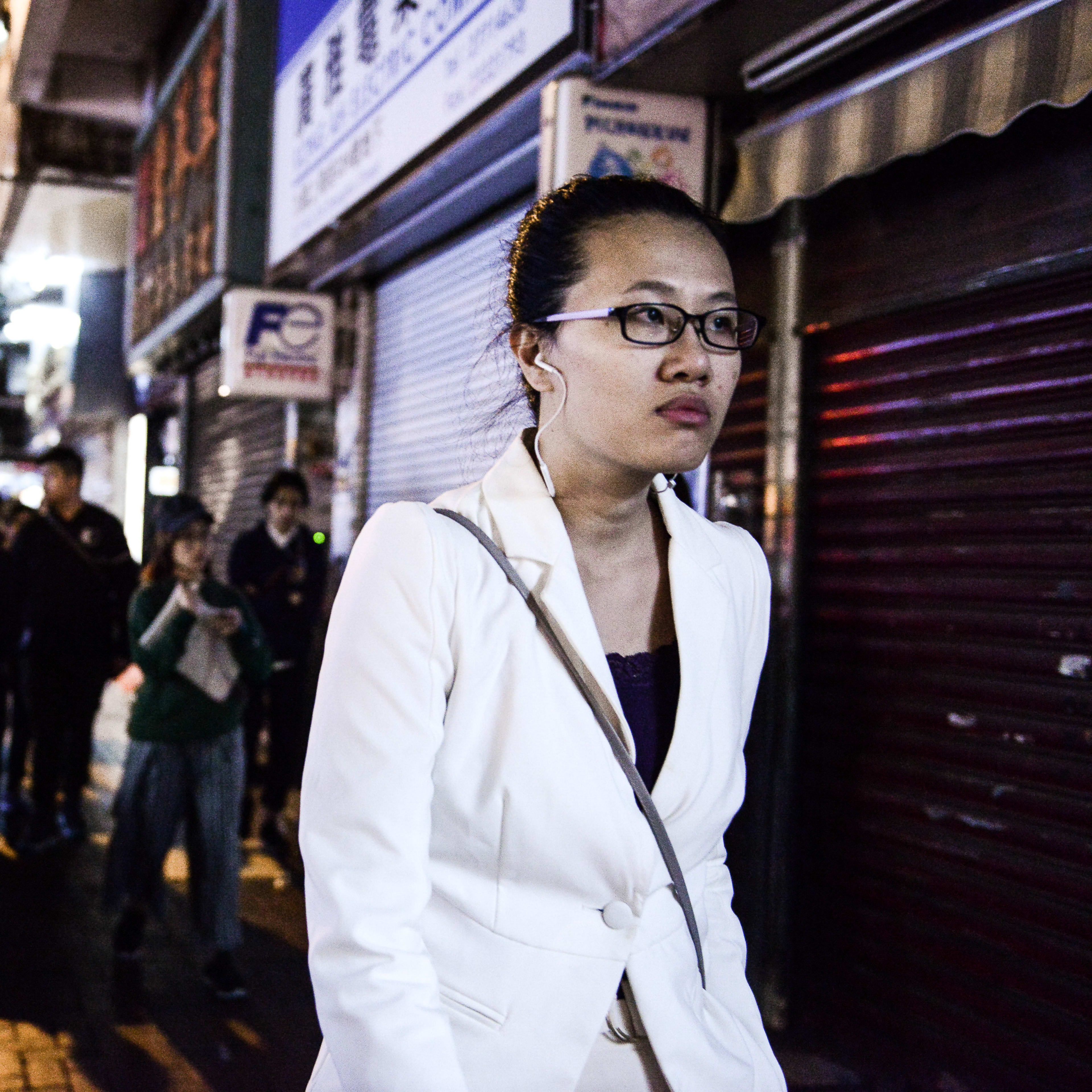 A candid, atmospheric street shot from Rooftop Productions’ contemporary Hong Kong theatre piece, Lost Shoreline (《消失的海岸線》). Actor Michelle Li is captured in the foreground, wearing a white blazer and earphones, walking through a dimly lit Hong Kong street at night. Her expression is thoughtful and detached, contrasting with the blurred figures of bystanders and a passing truck in the background, evoking a sense of urban isolation characteristic of the production. 'Lost Shoreline'