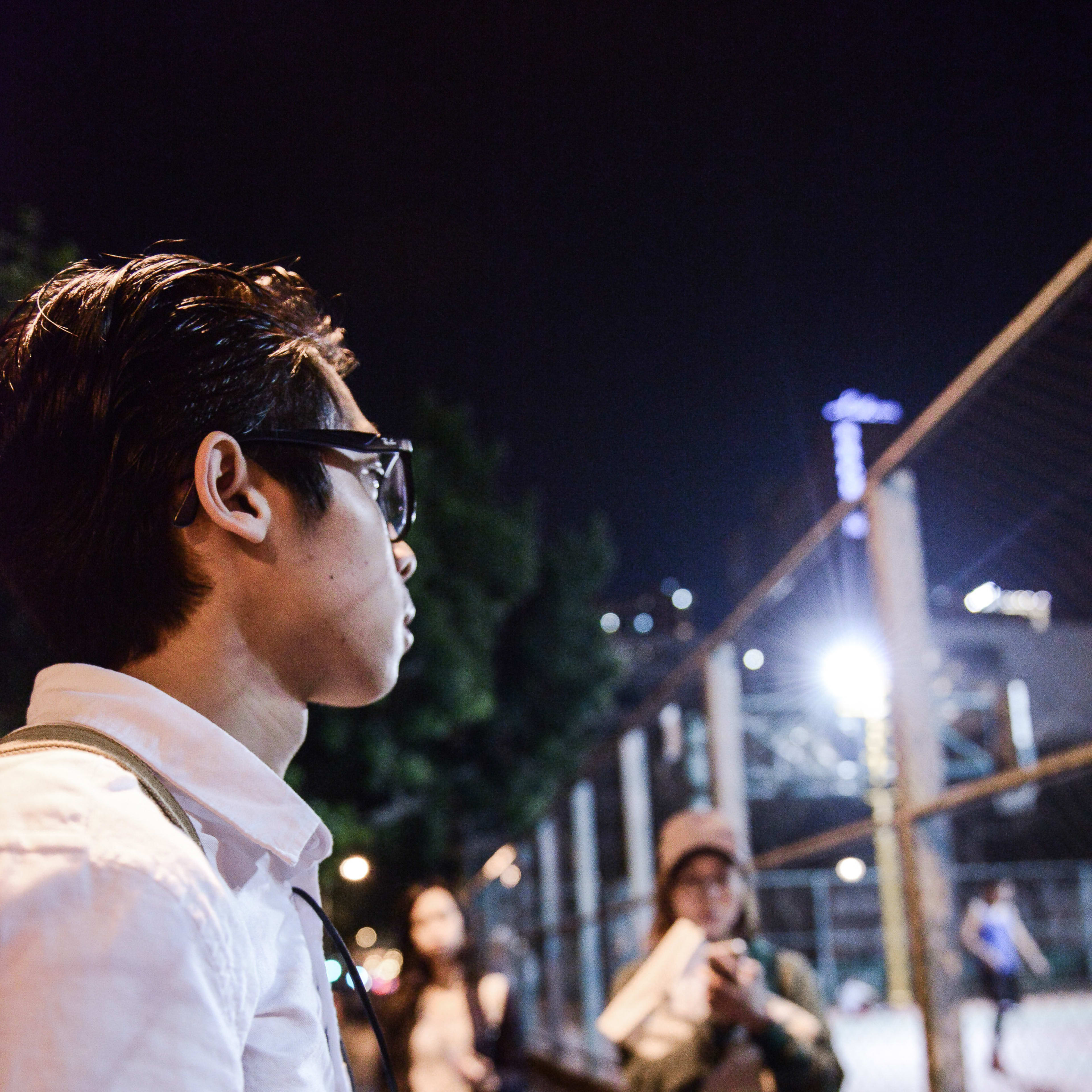 A behind-the-scenes rehearsal photo for the contemporary Hong Kong theatre production Lost Shoreline, featuring actor Chiu Chin Hei in profile, wearing a white shirt and glasses, holding a piece of rock in his palm while looking thoughtfully toward a lit outdoor court at night. In the background, other performers are blurred, capturing the contemplative atmosphere of this experimental stage performance by Rooftop Productions. 'Lost Shoreline'