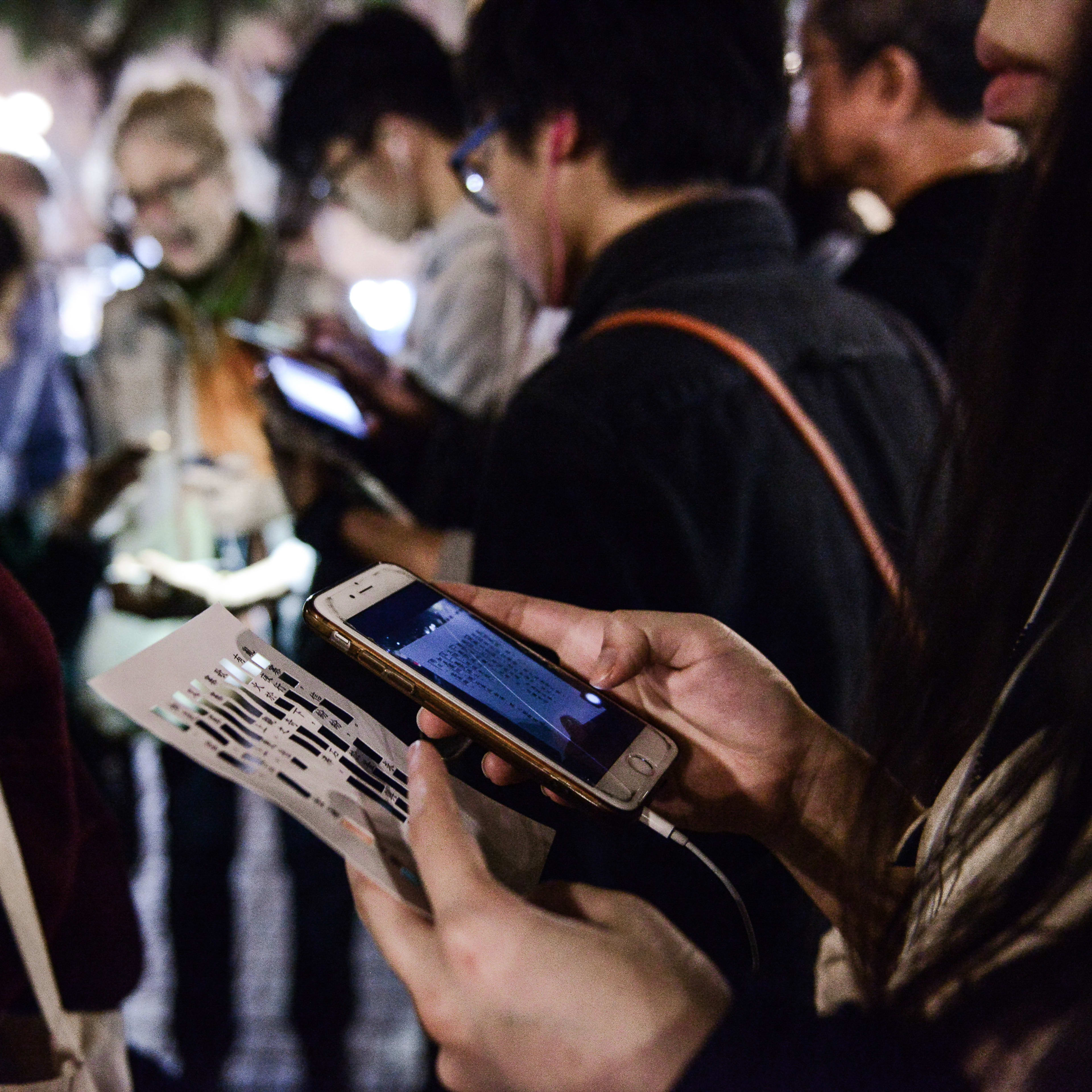 A close-up, dimly lit, atmospheric shot capturing the immersive experience of Rooftop Productions' contemporary theatre piece, 'Lost Shoreline'. A person in the foreground holds a mobile phone, using it to view a physical card or prop displaying text, reflecting the show’s innovative use of interactive technology and site-specific performance elements. Surrounded by other audience members focused on their devices, the scene highlights the intimate and participatory nature of HK contemporary theatre.