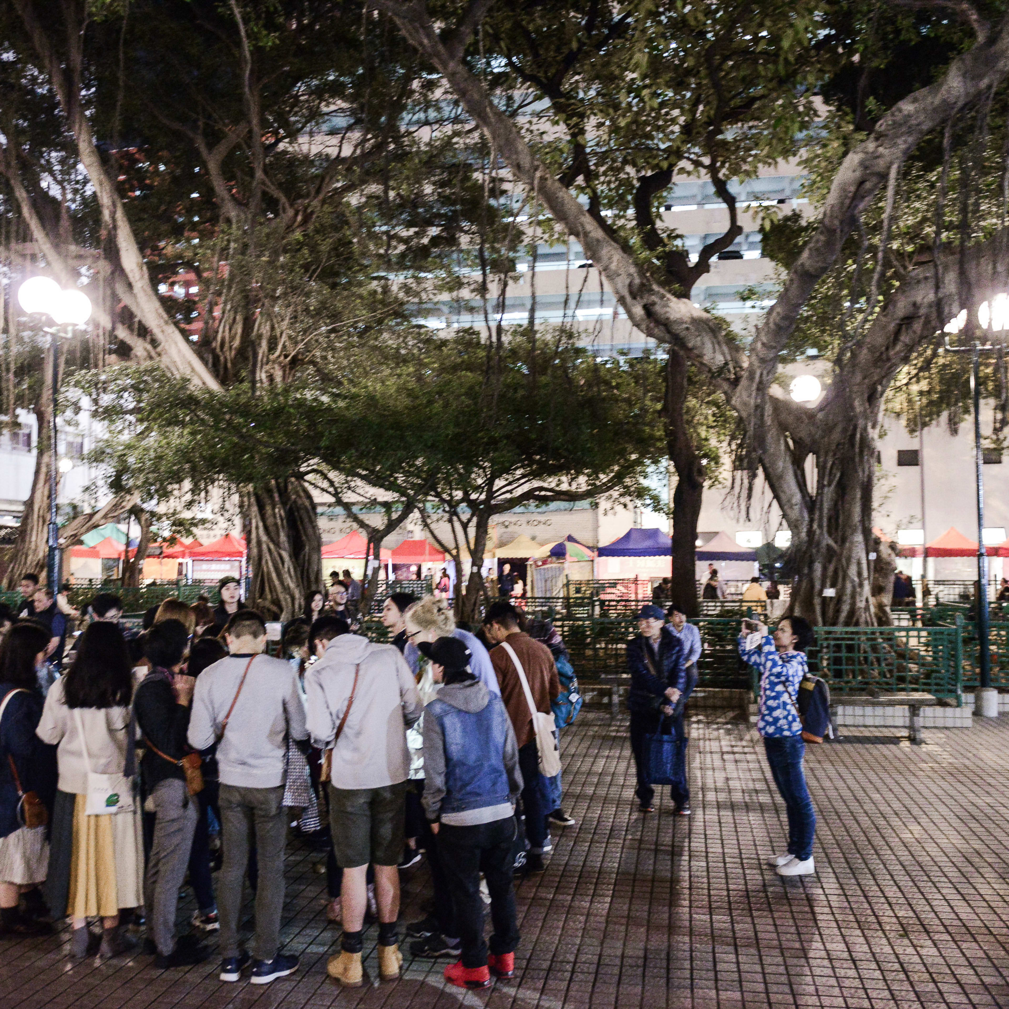 A wide-angle night shot of a workshop performance of 'Lost Shoreline' by Hong Kong’s Rooftop Productions. A diverse group of people gathers in a public plaza shaded by large banyan trees, illuminated by warm street lamps and colorful market tent stalls in the background. The participants are engaged in an immersive outdoor theatre experience, capturing the essence of contemporary HK community storytelling. 'Lost Shoreline'