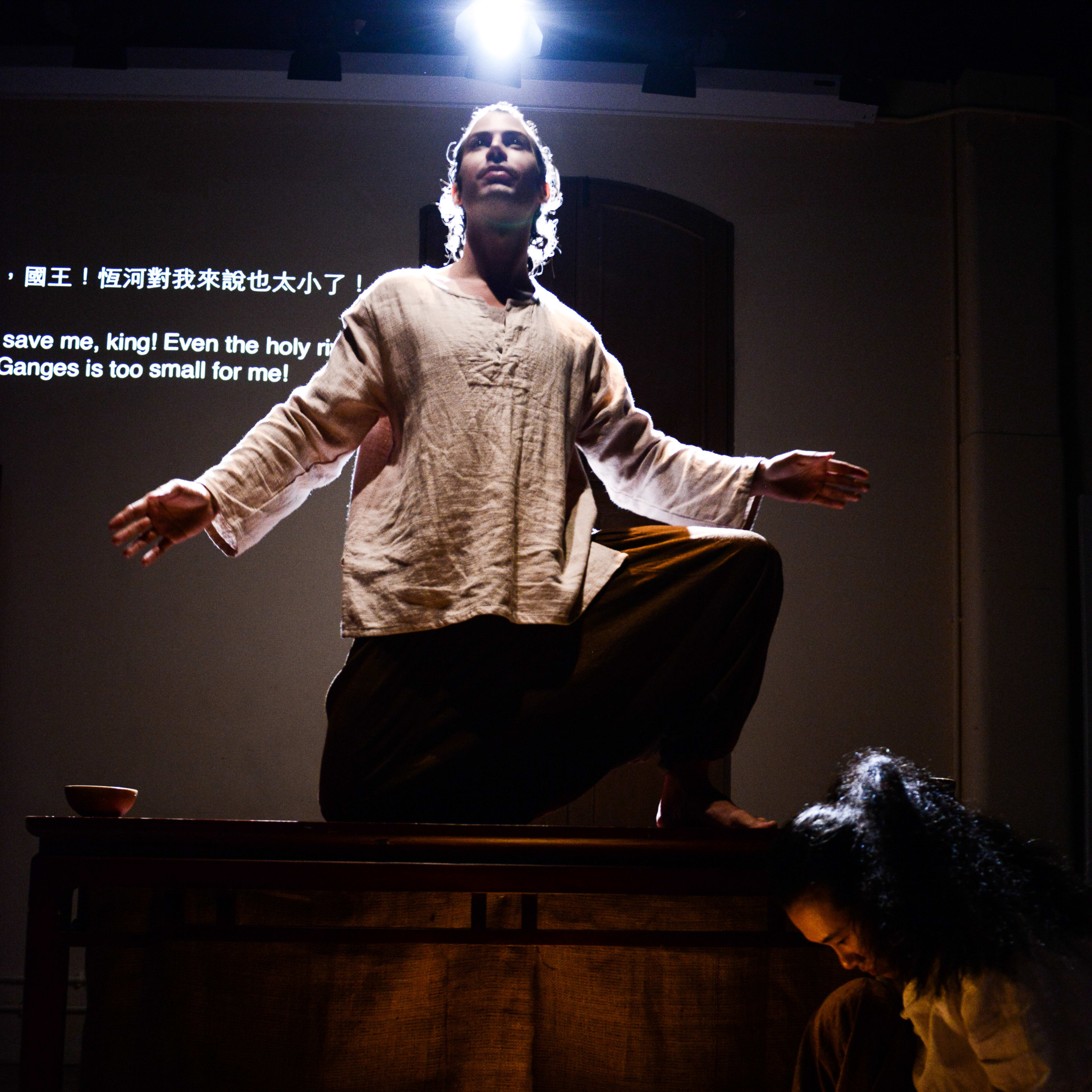 在天台製作的當代劇場作品《牛奶與蜜糖》中，演員艾浩家（Ivor Houlker）站在木桌上，雙臂向外延伸，呈現出一種神聖激昂的姿態。在他下方，另一位表演者在舞台地板上一盞發光的缽旁跪地沉思。背景投影上的字幕寫著：「國王！恆河對我來說也太小了！」
