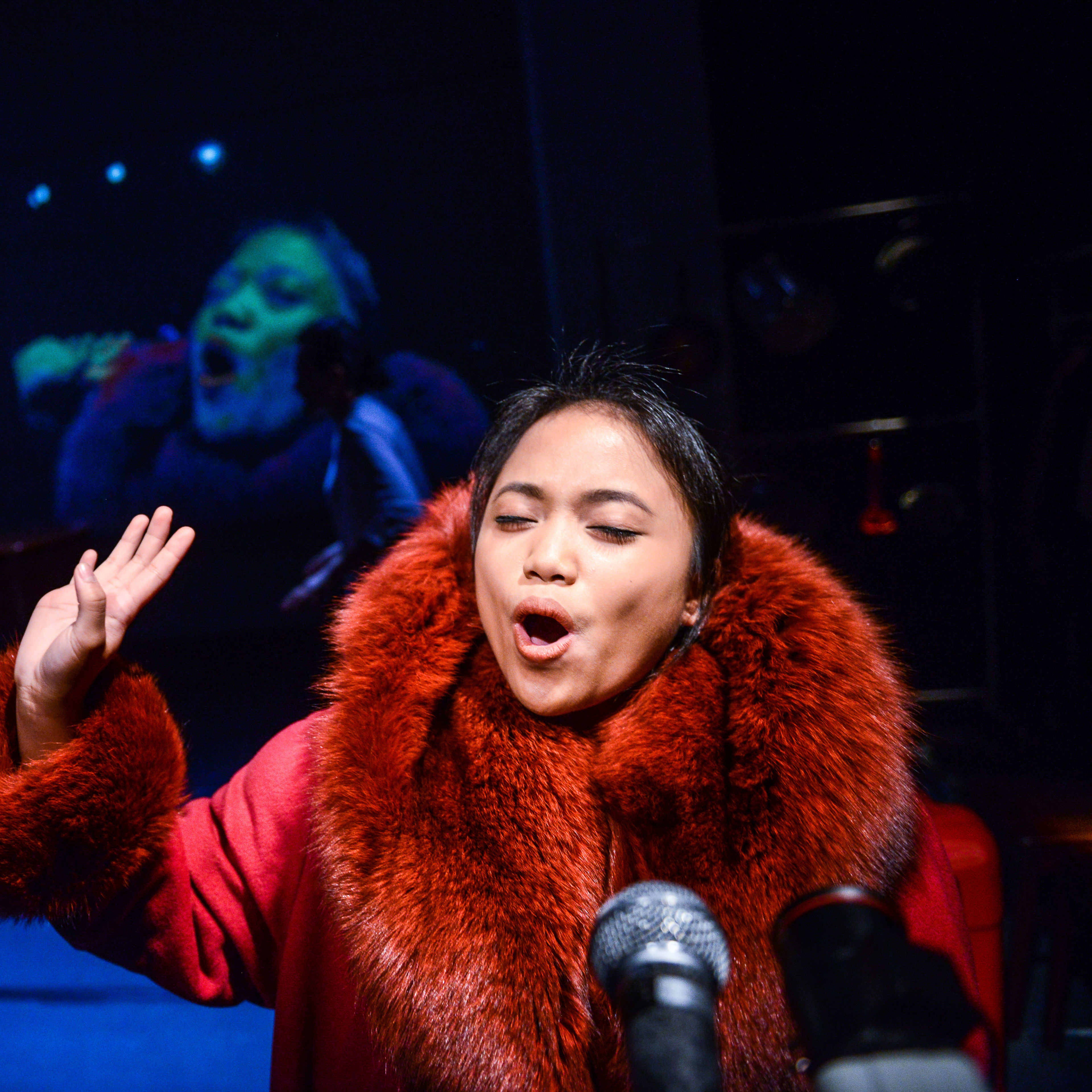 演員區洛思 (Russell Terre Aranza) 在天台製作 (Rooftop Productions) 的當代香港劇作《不是女僕》中登台演出。在冷調藍色燈光的映襯下，她身穿帶有誇張毛領的紅色大衣，神情專注且動作豐富，背景投射出一張巨大的變形人臉，營造出強烈的戲劇氛圍。區洛思在台上演繹道：「你們在做什麼？我看開會回來，而我期望所有東西都完美。」