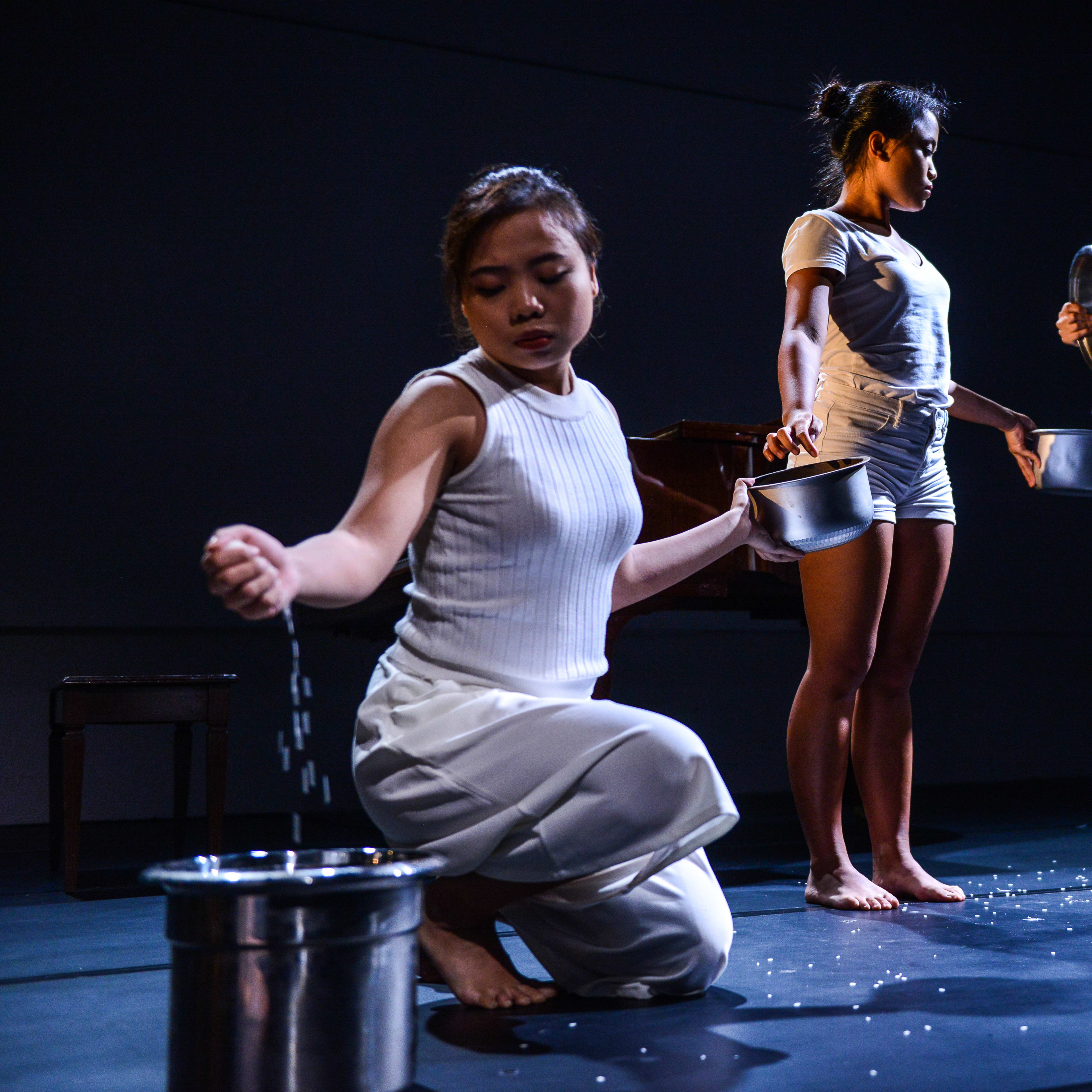 三位表演者身穿純白服飾，站立在天台製作（Rooftop Productions）當代劇場作品《不是女僕》(Not The Maids) 的極簡黑色舞台上。前景的表演者跪地，將手中的顆粒緩緩灑入金屬桶中，後方的兩位表演者則站立著，將盆中的顆粒傾倒而下。戲劇性的高對比燈光突顯了這種重複且具有儀式感的動作，成為該劇探討家務勞動議題的強烈視覺隱喻。此場景呈現了「全球化親職外判鏈」。