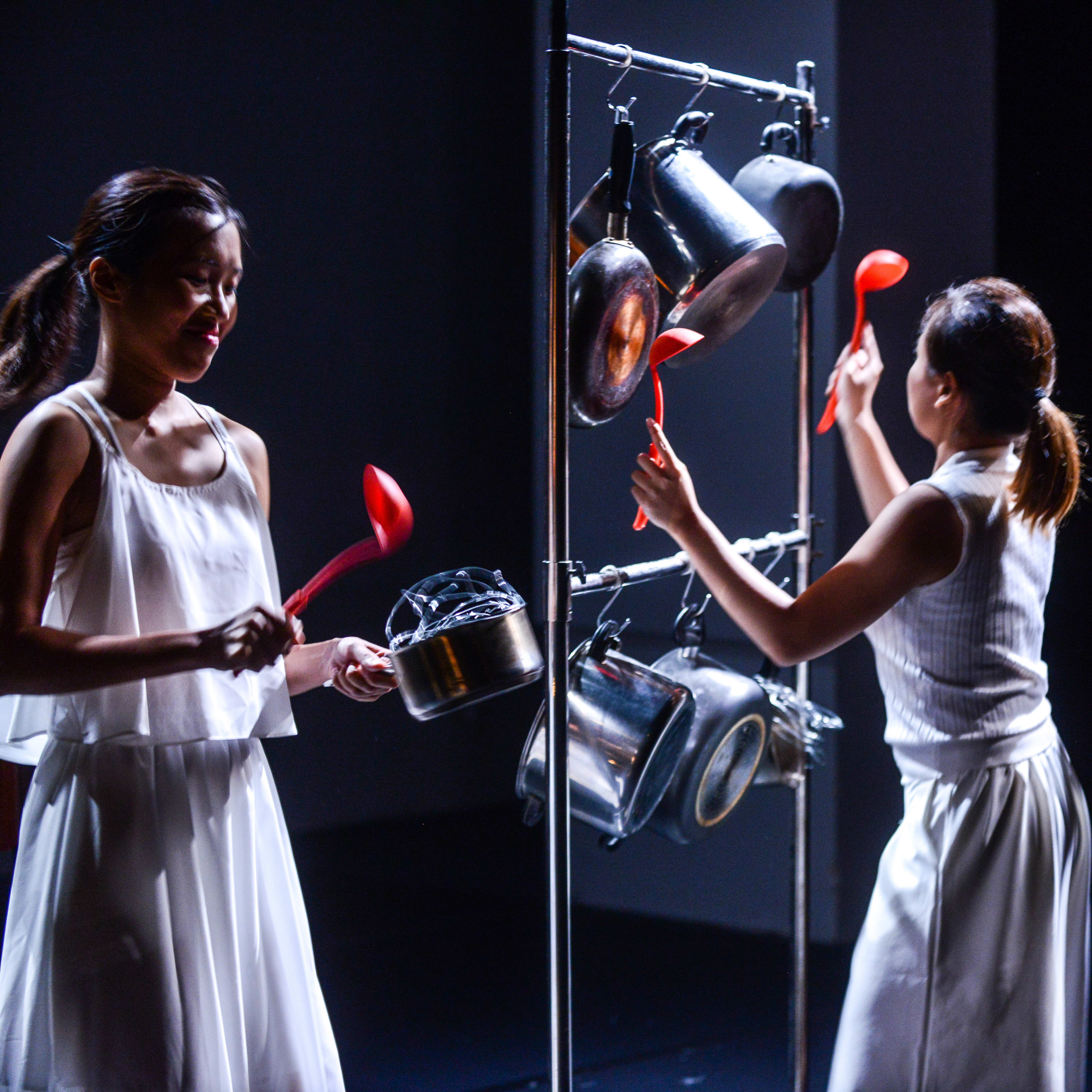 三位穿著白色服裝的演員在天台製作 (Rooftop Productions) 的當代香港劇場作品《不是女僕》(Not The Maids) 排練現場。舞台燈光陰暗且充滿戲劇張力，演員們手持鮮紅色的湯勺，有節奏地敲擊懸掛在金屬架上的鍋具，以敲擊樂呈現出家庭生活中的緊繃感。「如果她在半夜洗碗，便會吵醒我。」