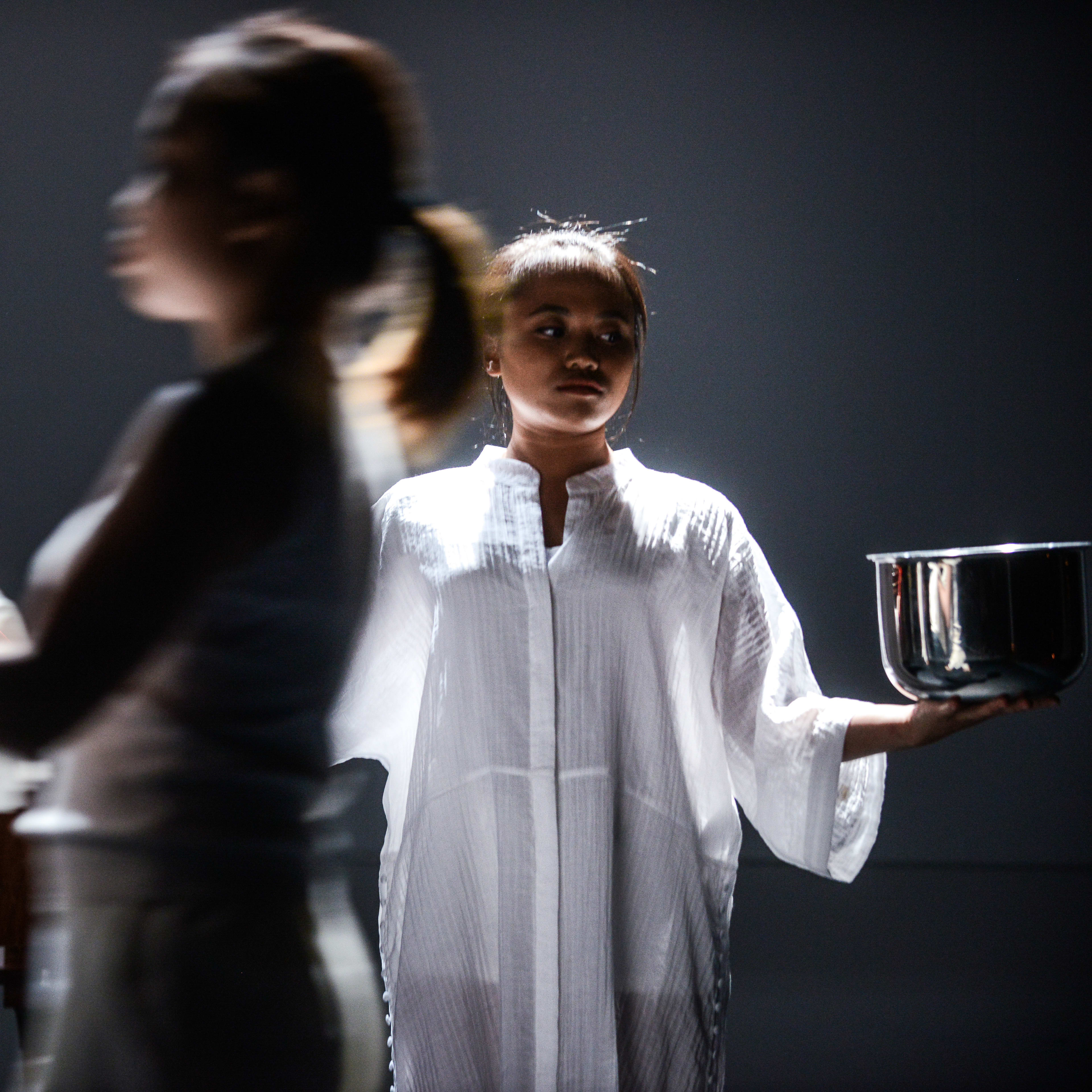 在這張由「天台製作」（Rooftop Productions）呈獻的當代香港劇場作品《不是女僕》劇照中，燈光營造出充滿戲劇張力的陰影氛圍。一位演員清晰地立於舞台中央，手中持有一個金屬鍋；前景中另一位演員的身影略顯模糊，正捧著一塊白布與一顆紅色物件，為畫面增添了儀式感與緊張情緒。此影像捕捉了《不是女僕》極簡而充滿張力的舞台風格。區洛思深情演繹台詞：「聆聽我們，上主，請好憐憫，因我們對您所犯的罪。」