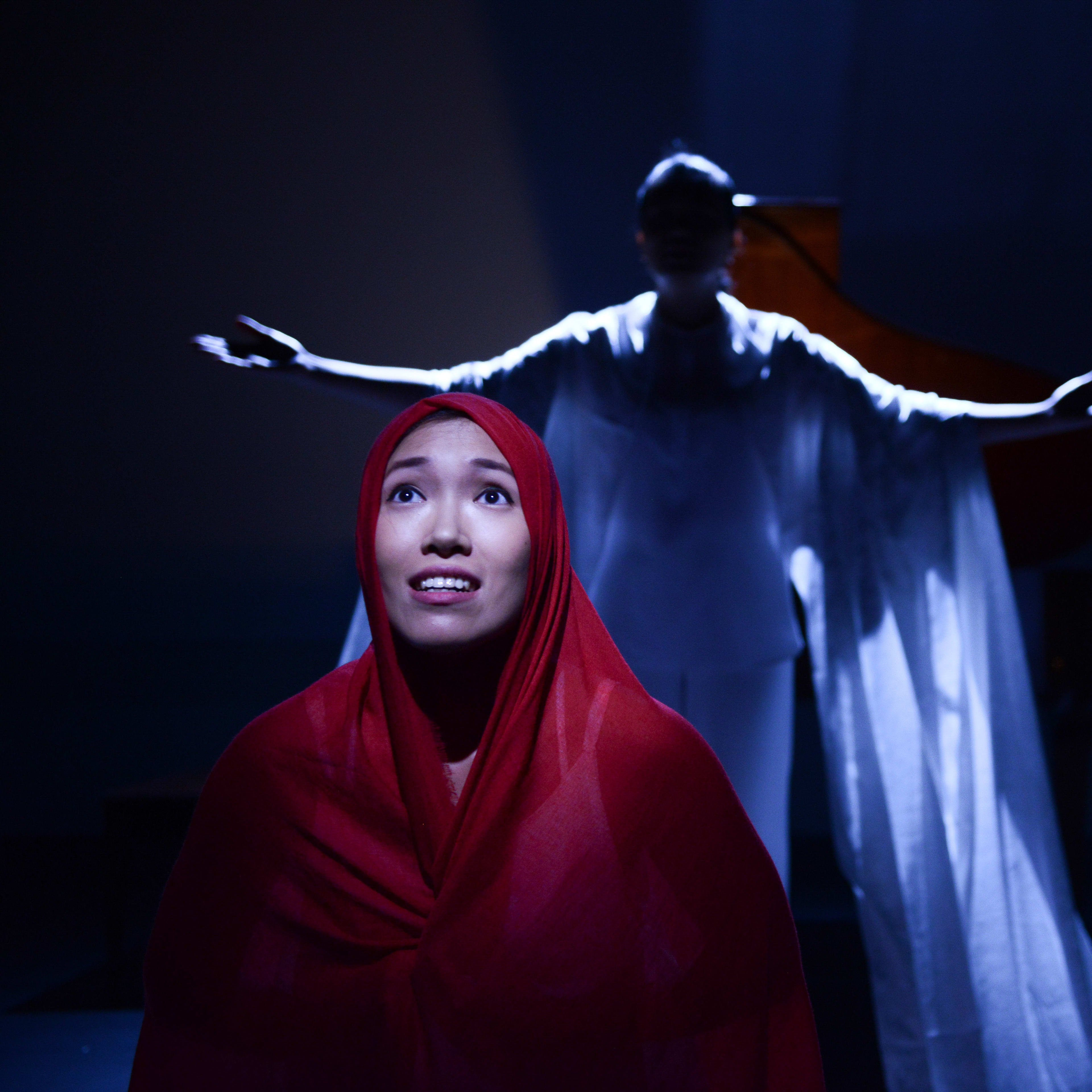 在天台製作 (Rooftop Productions) 的當代劇場作品《不是女僕》(Not The Maids) 中，演員梁皓貽（Isabella Leung）身披紅色頭巾位於畫面中央，表情中流露著深沉的渴望與脆弱。背景中，另一位表演者在冷冽的藍色舞台燈光下，雙臂向兩側伸展，呈現出莊嚴且具儀式感的姿態，右側則有另一名手持物件的表演者。極簡的舞美設計深刻體現了這部香港當代劇場作品的情感張力。表演者訴說著劇中的核心提問：「為什麼外傭必須要留宿於僱主家中？」