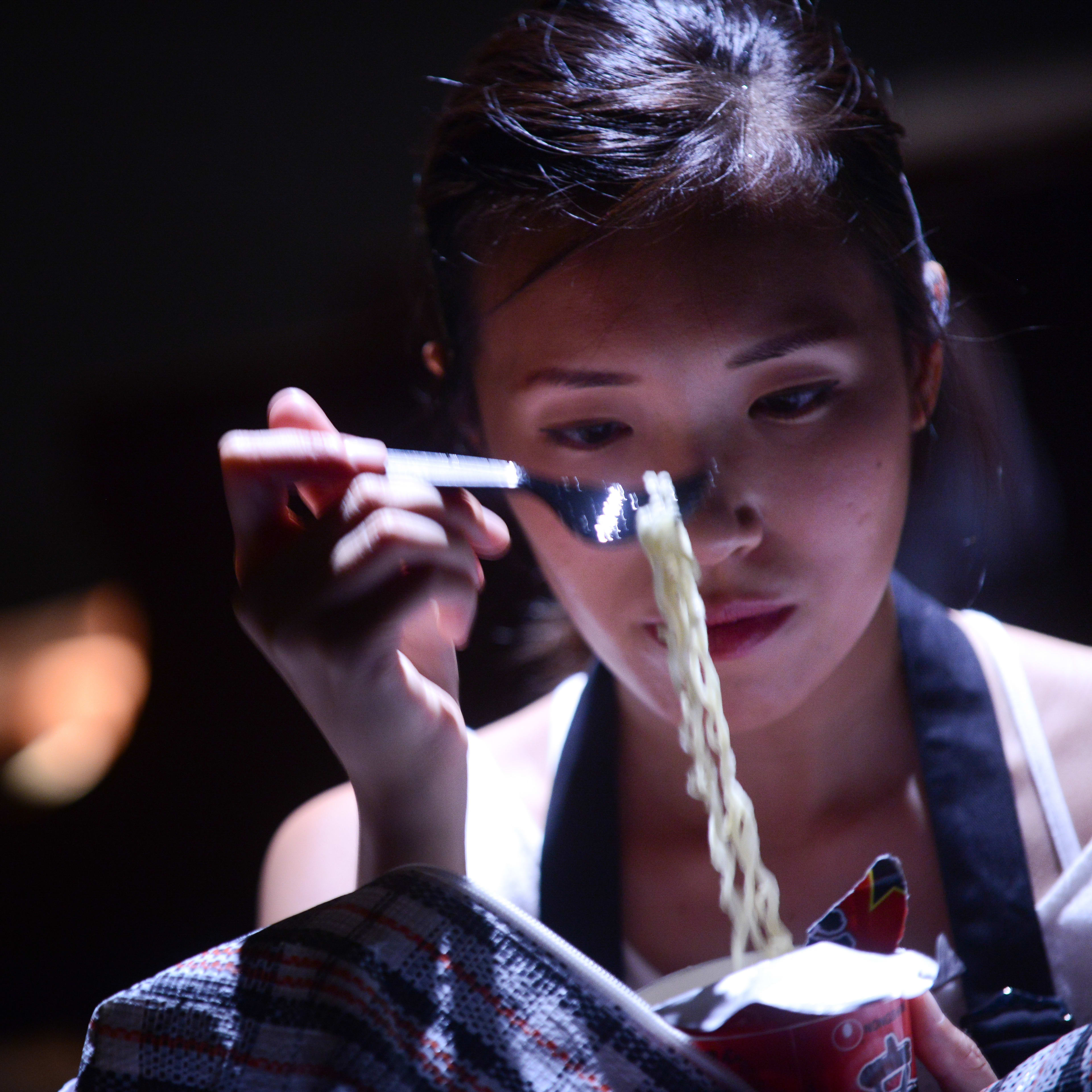 在這張來自天台製作（Rooftop Productions）當代香港戲劇《不是女僕》的劇照中，演員梁皓貽（Isabella Leung）置身於昏暗的舞台燈光下，展現出充滿張力的戲劇時刻。她神情專注地拿著一杯杯麵，用叉子挑起麵條，陰影下的光影勾勒出她沉思的神情，深刻呈現了演出中那份貼近日常的真實感。當角色回憶起這段相處時，她說道：「他知道我有一些杯麵，然後他會拿來吃。」