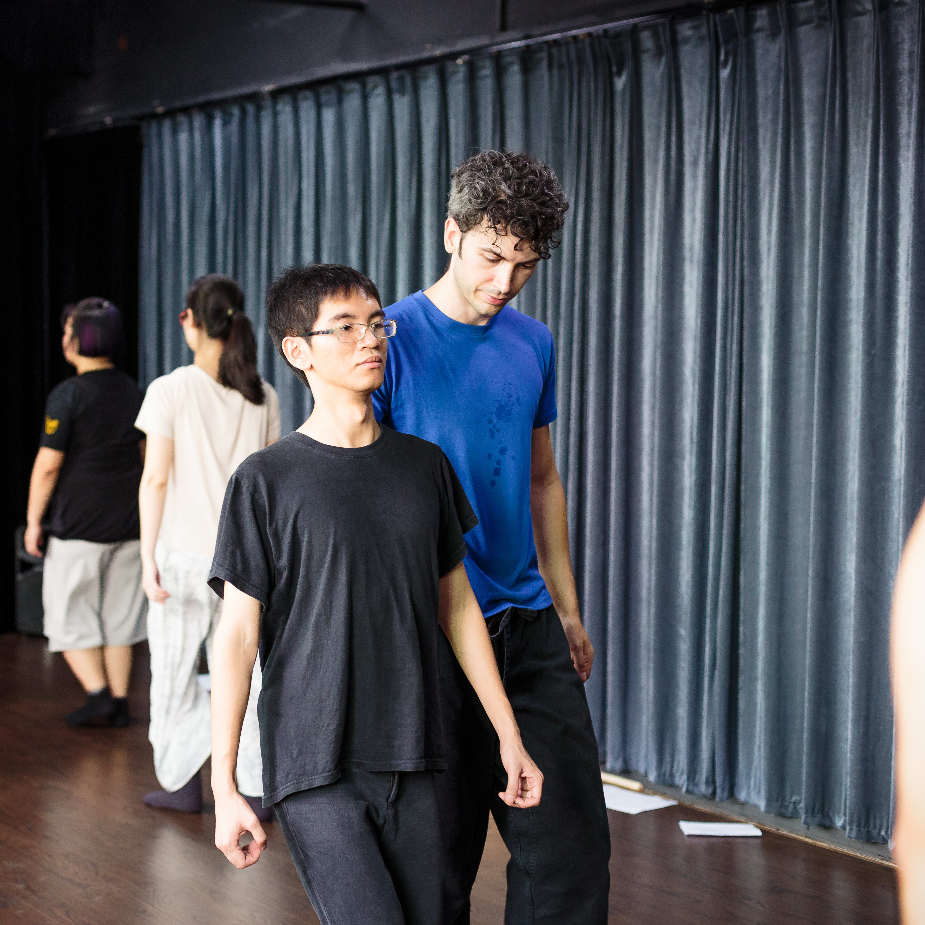 學員於「廣州形體劇場及復調演唱工作坊」中進行專注的肢體訓練，此活動為天台製作（Rooftop Productions）推動的當代劇場藝術交流。前景中，導師正帶領學員練習形體劇場的動作，背景中的表演者亦投入於訓練中，展現了當代香港劇場對身體表達與敘事的重視。「廣州形體劇場及復調演唱工作坊」