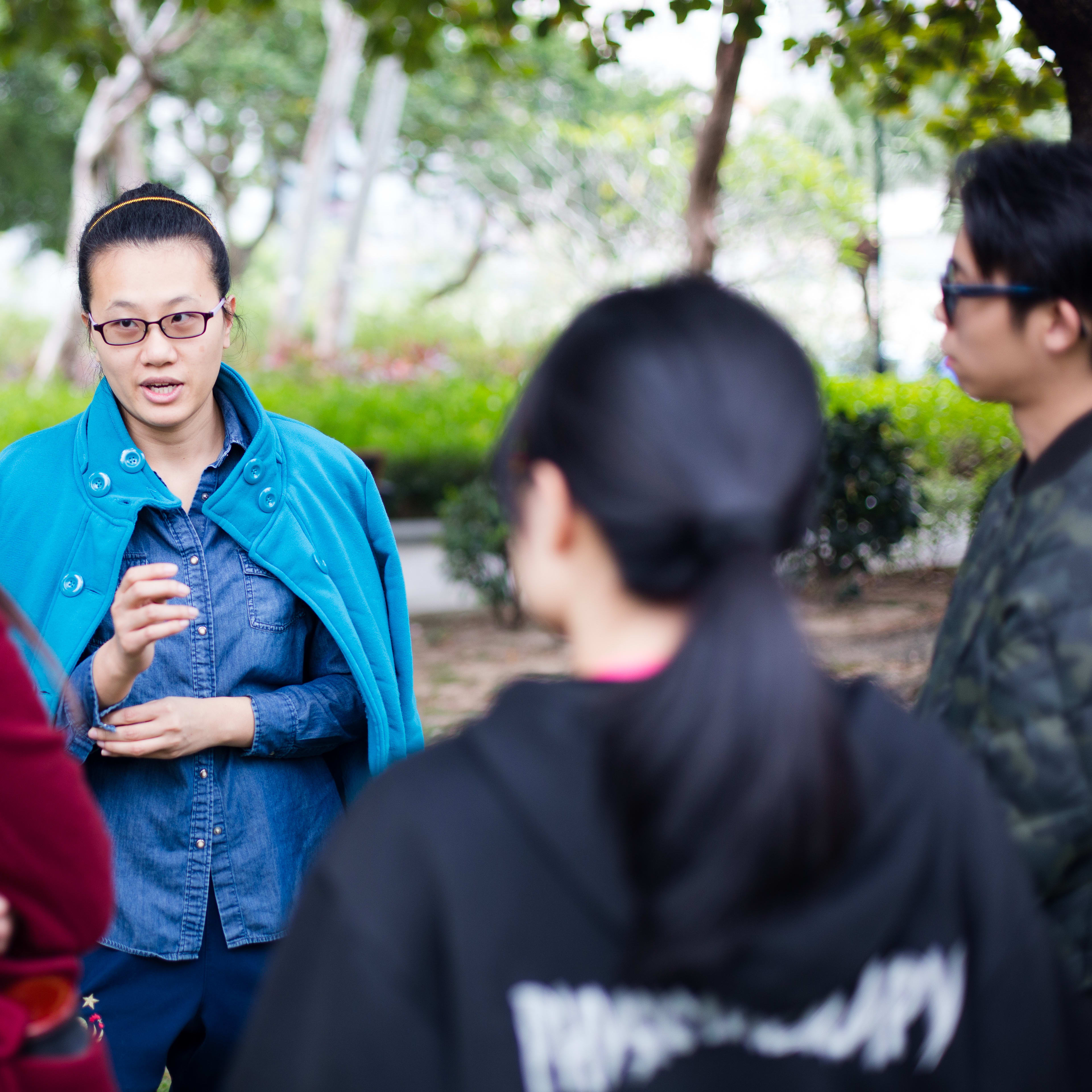 在戶外公園的場景中，藝術家李婉晶（Michelle Li）正與參與者進行深入的藝術交流。這是「歐洲形體劇場實踐工作坊」（Polyphonic Song: Voice and Theatre Practice）的精彩時刻，由天台製作（Rooftop Productions）策劃，旨在探索聲線訓練與當代劇場實踐的交匯點，致力推動香港舞台藝術的發展。「Song Theatre」