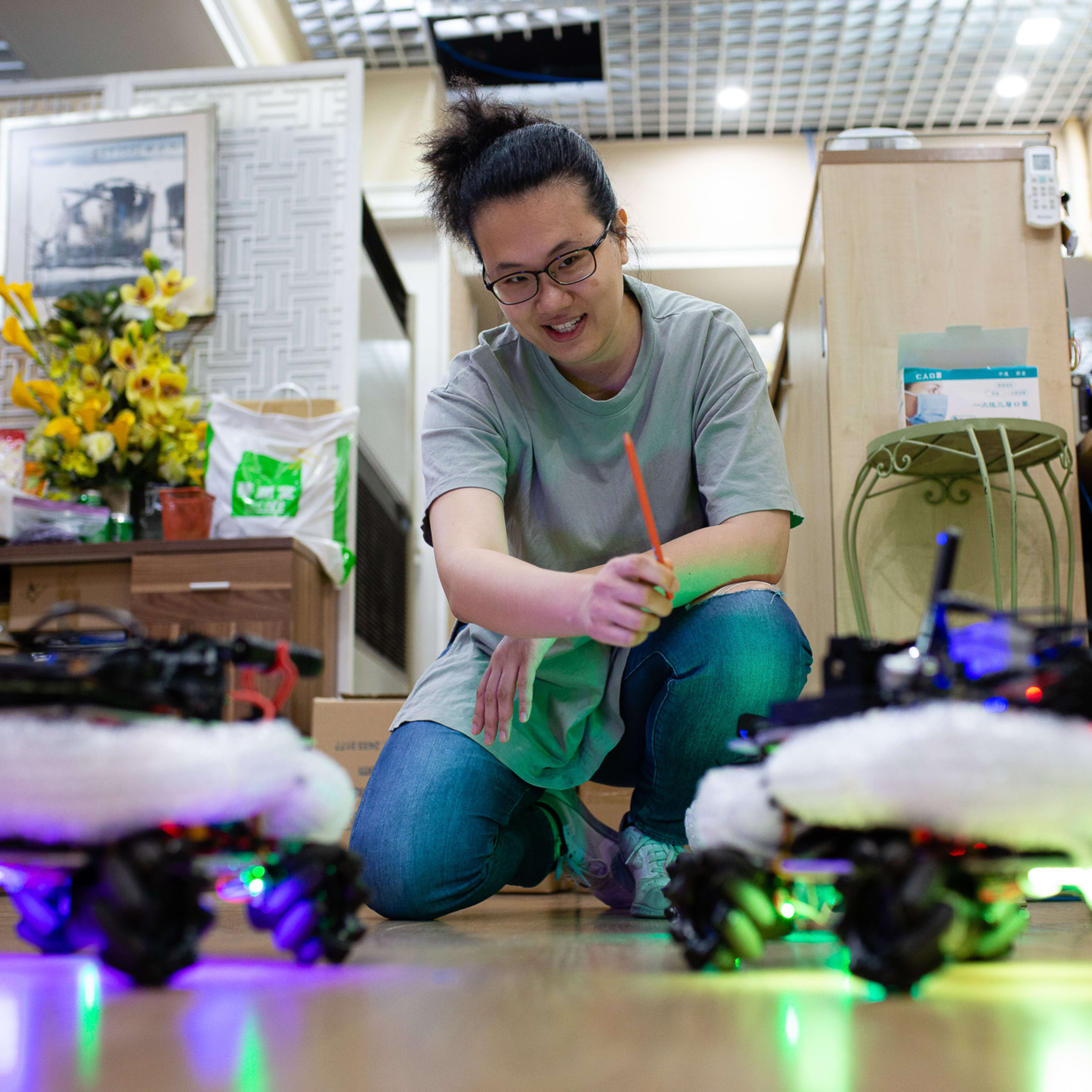 In a behind-the-scenes rehearsal for the Rooftop Productions contemporary theatre piece 'Presence' (遙距在場), performer Michelle Li kneels on the floor, smiling as she holds a pen while interacting with two small, illuminated toy robots featuring omni-directional wheels. The warm, dimly lit room is filled with creative props, reflecting the experimental and DIY nature of Hong Kong's independent theatre scene. As Michelle remarks during the rehearsal, 'Even the pens are salvaged.'
