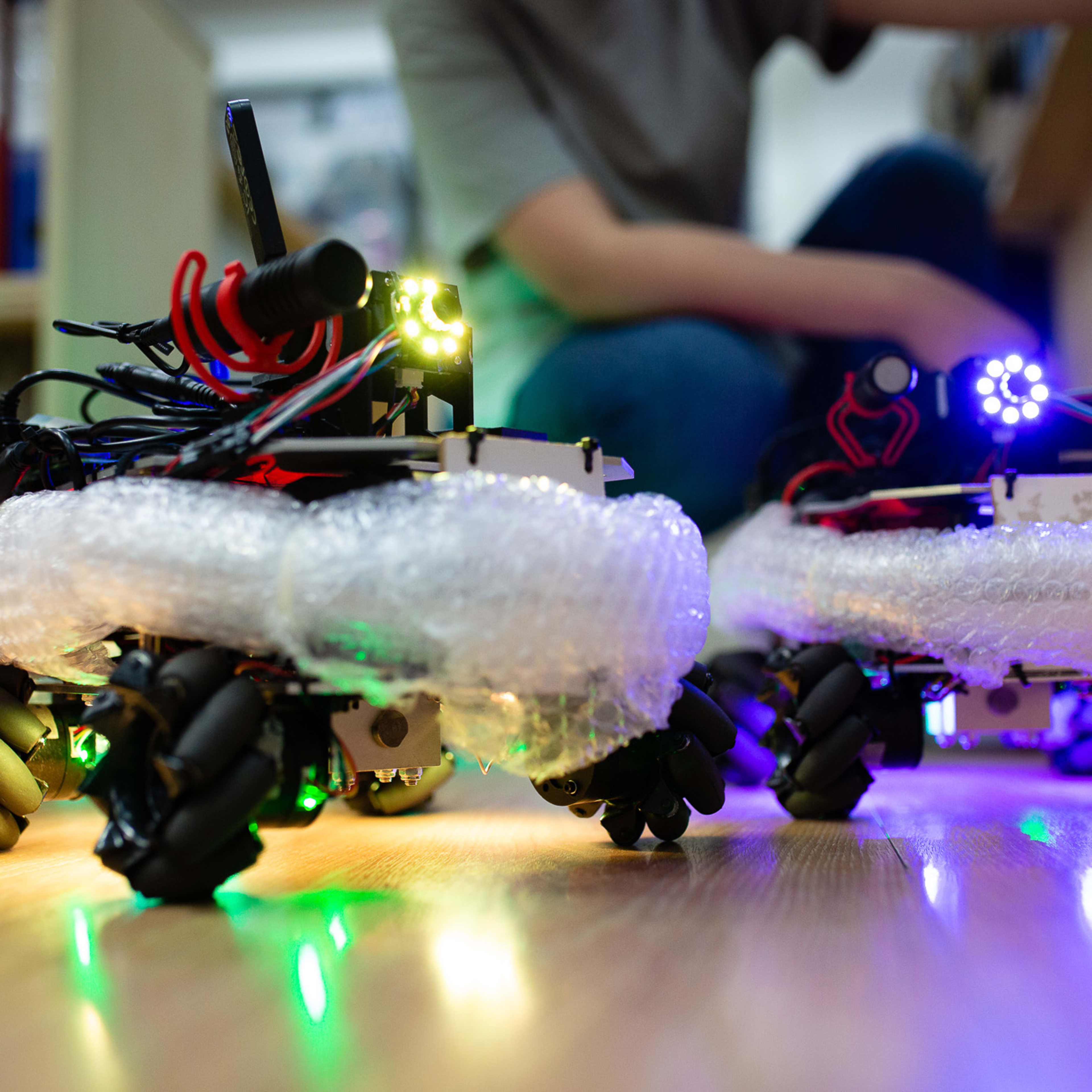 Two small, robotic vehicles equipped with Mecanum wheels and glowing LED lights navigate a wooden floor, surrounded by bubble wrap, as part of a creative rehearsal for the contemporary Hong Kong theatre production 'Presence' by Rooftop Productions. In the soft-focus background, a person works at a desk, capturing the experimental spirit of this performance piece. 'Around the office.'