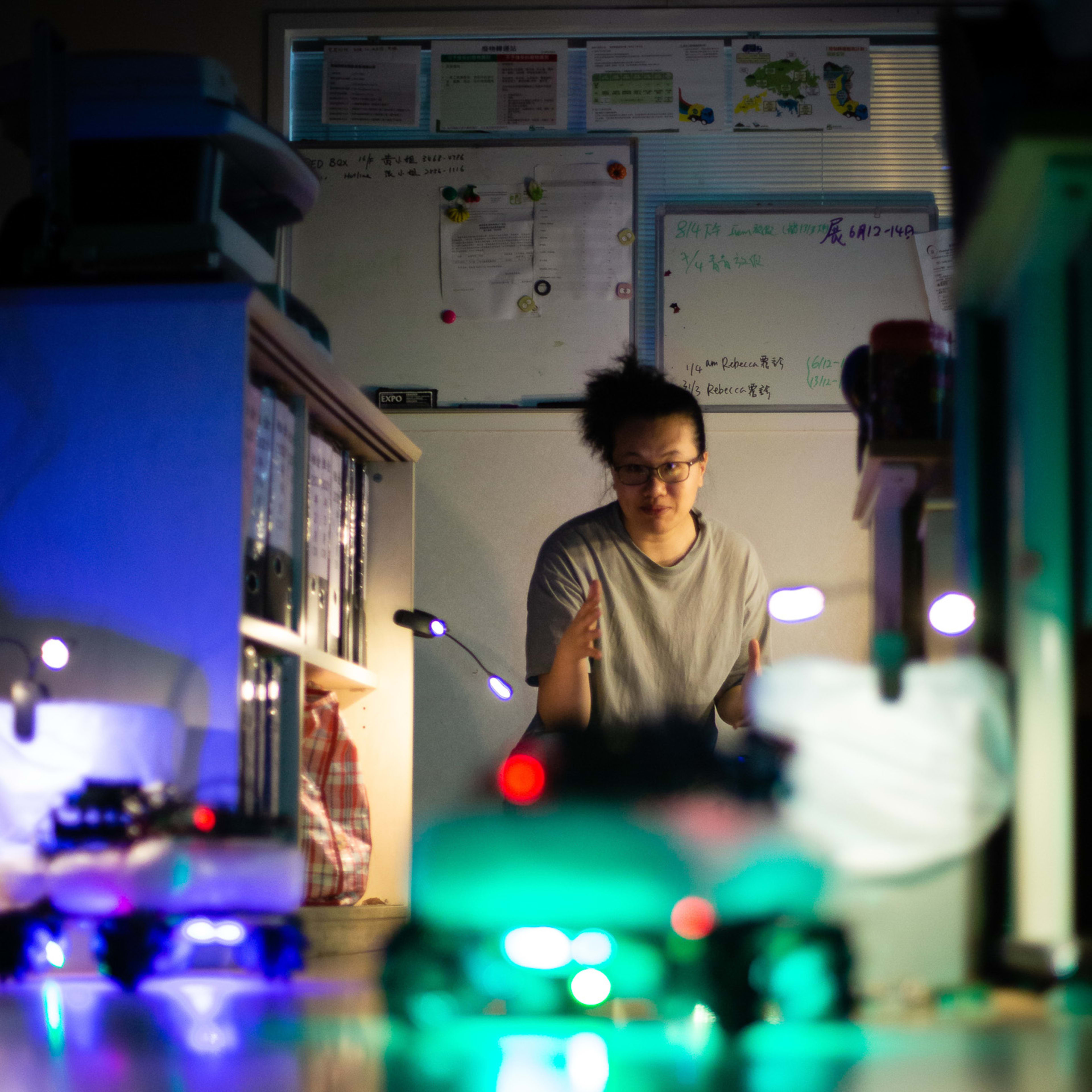 In a rehearsal for Rooftop Productions' contemporary theatre piece 'Presence' (遙距在場), performer Michelle Li (李婉晶) sits in a dim office setting, illuminated by the bright, blurred green light of a small robot in the foreground. The atmospheric lighting creates a sense of mystery and technological intimacy. Michelle gazes forward with a focused, contemplative expression, her hands gesturing as if she is navigating the space. This moment captures her saying, "Second task: Find the keys."