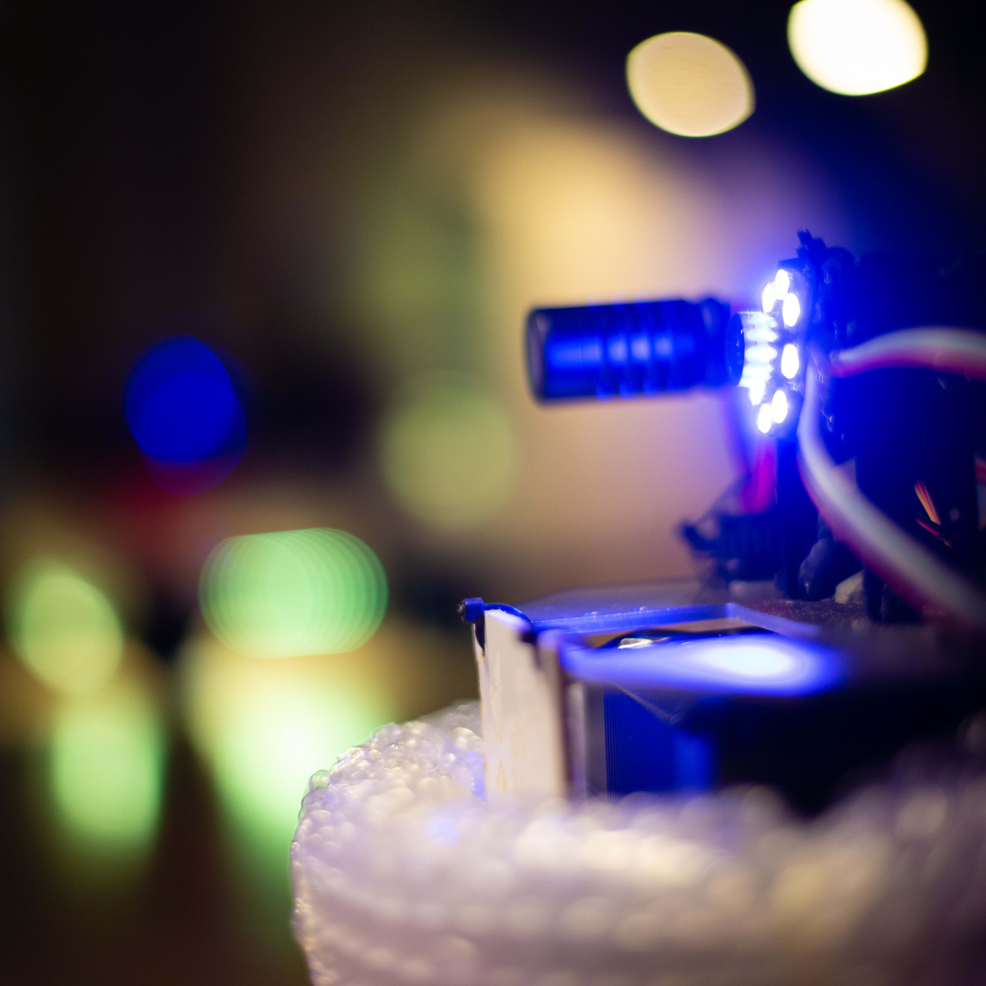 A close-up, atmospheric shot captures a small, high-tech robot assembly used in Rooftop Productions' contemporary theatre piece, 'Presence' (《遙距在場》). The focal point is a camera lens and microphone rig illuminated by a striking blue LED ring, contrasting with a soft, bokeh-blurred background. This device serves as the performative 'eye' in the production, highlighting the intersection of technology and live performance in Hong Kong's experimental theatre scene. 'Close up of the car's camera and mic'