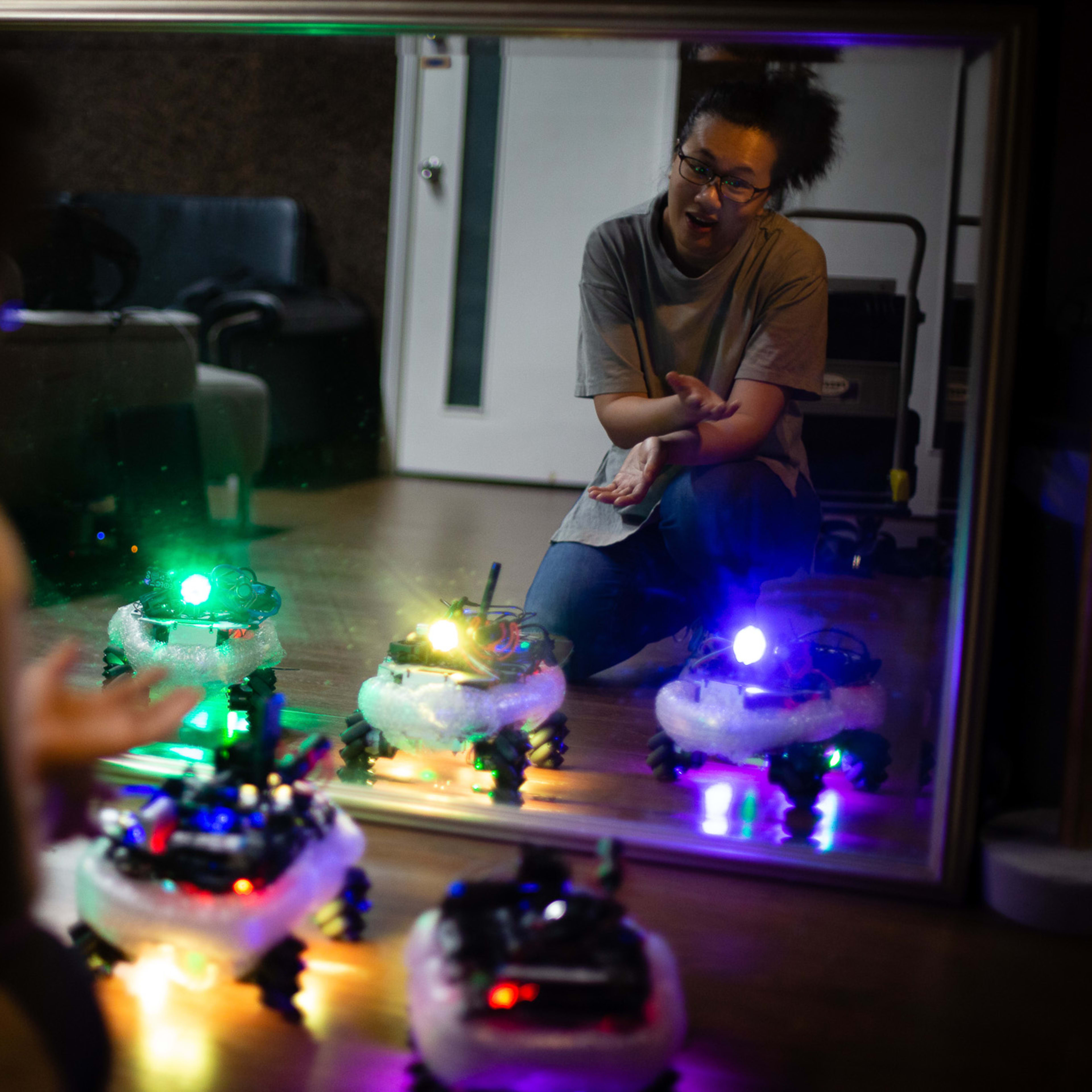 In a rehearsal for the contemporary Hong Kong theatre production 'Presence' (《遙距在場》), actor Michelle Li sits on the floor, interacting with a small, brightly lit remote-controlled robot. The scene is captured through a mirror reflection, creating a layered, intimate aesthetic that plays with themes of distance and digital connection. Michelle, focused on the device, gestures as she follows instructions. The robot glows with vivid green and purple LED lights, highlighting the technological elements often found in Rooftop Productions' avant-garde stage works. Michelle Li says, "FIND POST-ITS, FORM A SENTENCE AND INFORM COLLEAGUE IN ROOM."