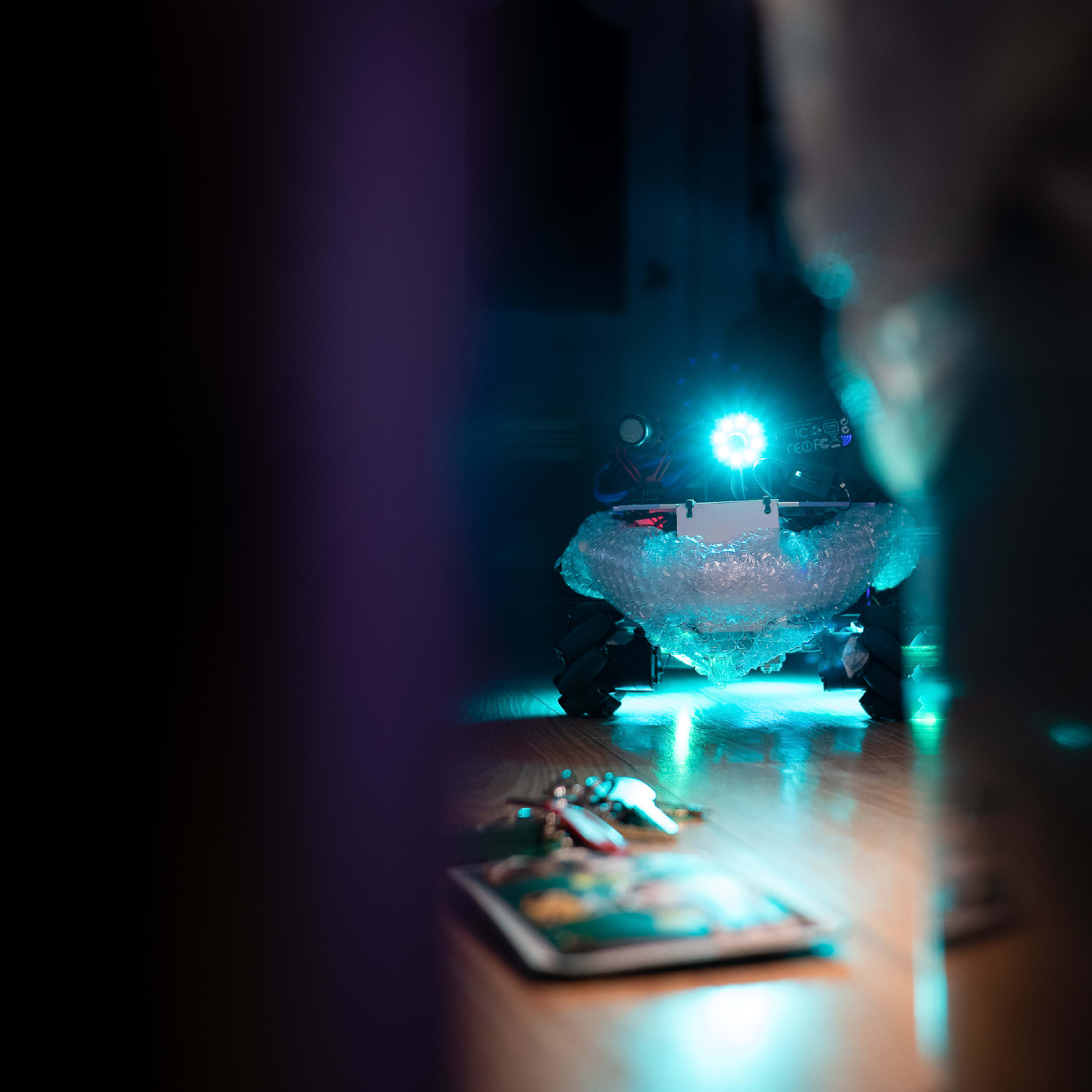 A close-up shot from the Rooftop Productions (天台製作) experimental performance 'Presence' (《遙距在場》), featuring a small, remote-controlled robot wrapped in bubble wrap illuminated by a glowing blue LED light in a dimly lit room. In the foreground, a set of keys rests on a wooden floor, highlighting the intersection of technology and intimacy in contemporary Hong Kong theatre. As the performer guides the device, they reflect: "Completed the second task: find the keys."