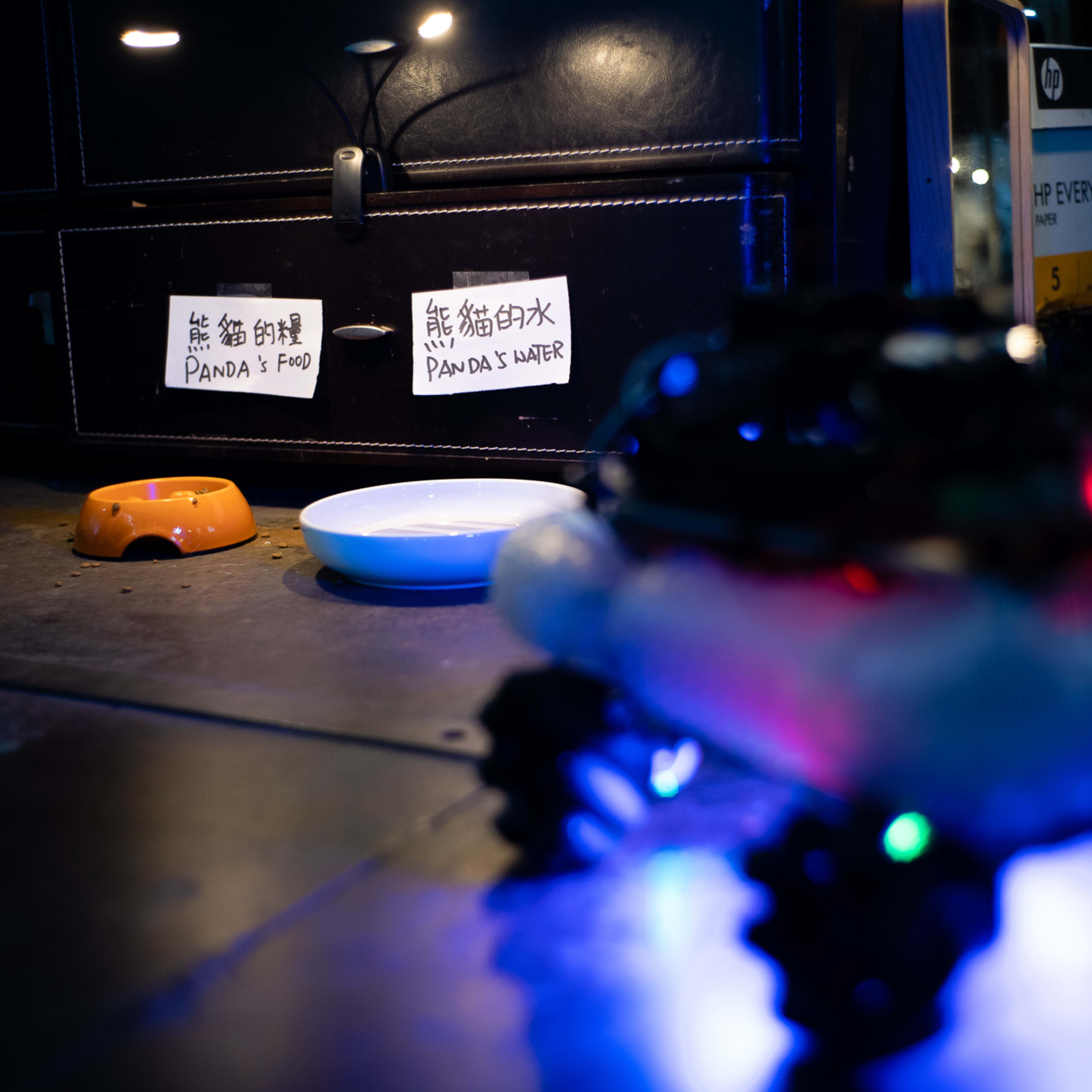 A close-up shot from the Rooftop Productions (天台製作) experimental performance 'Presence' (《遙距在場》), featuring a low-angle view of a dimly lit stage floor. Two white labels on a dark drawer front read 'Panda’s Food' and 'Panda’s Water,' positioned above a small orange food bowl and a shallow white water dish. In the foreground, a robotic device with glowing blue and red LED lights blurs out, emphasizing the high-tech, minimalist aesthetic of this contemporary Hong Kong theatrical production. 'Panda's bowl and water'