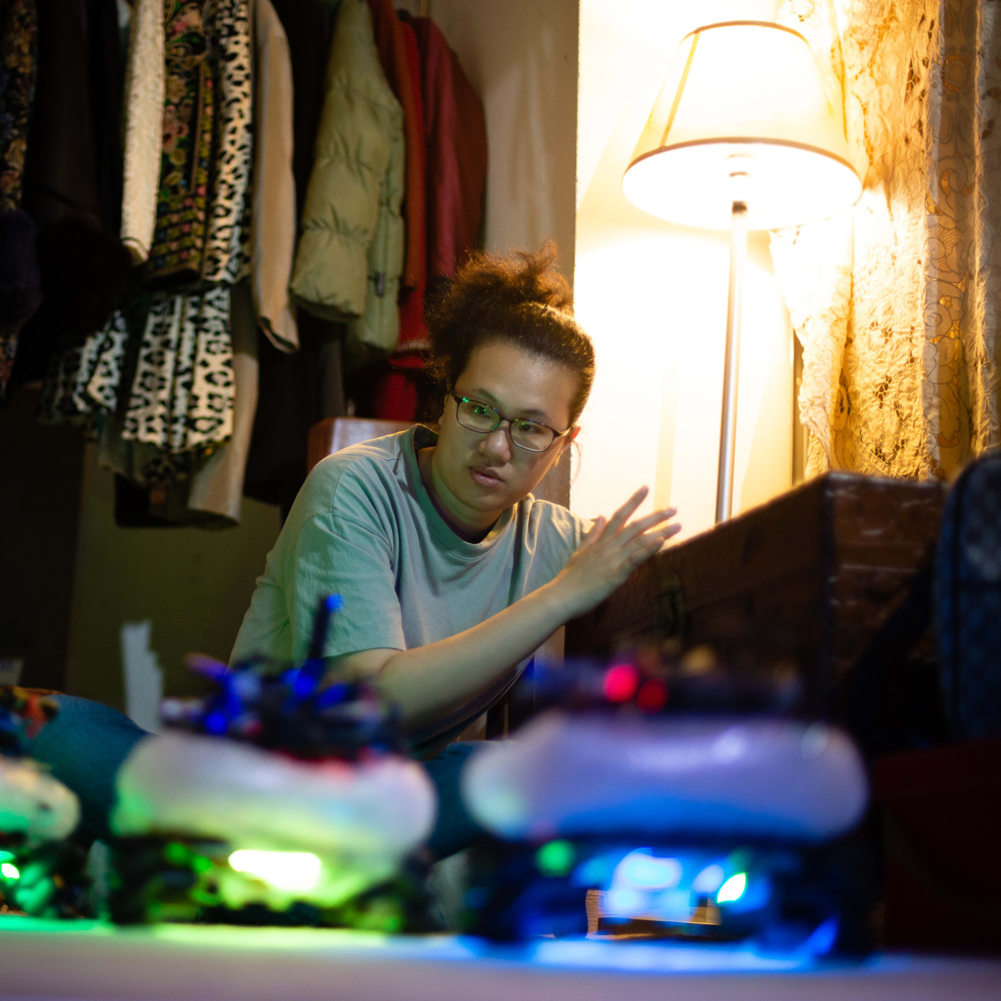 In a poignant rehearsal scene from Rooftop Productions’ contemporary Hong Kong theatre piece 'Presence' (《遙距在場》), actor Michelle Li sits on the floor of a dimly lit room filled with hanging garments. She gestures thoughtfully towards a traditional camphor wood chest, illuminated by the vibrant, neon-colored glows of small robotic devices at her feet. This intimate moment highlights the interplay between technology and memory in experimental stage performance. As she interacts with the props, she reflects: 'The camphor wood chest'.