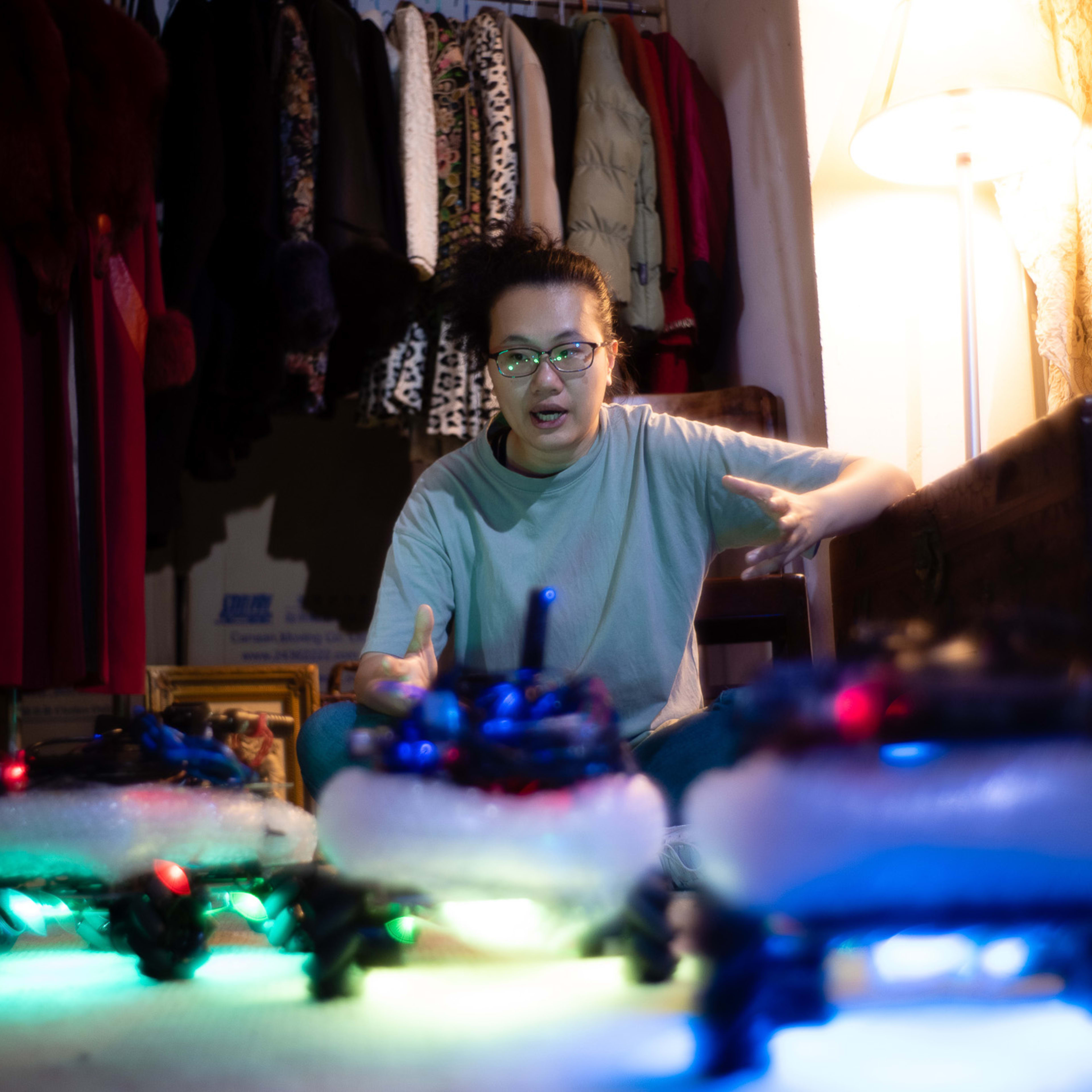 In this rehearsal photo for Rooftop Productions' (天台製作) contemporary theatre piece 'Presence' (《遙距在場》), performer Michelle Li sits on the floor surrounded by three small, robotic devices emitting vibrant green and blue LED lights. Li, wearing glasses and a light-colored t-shirt, gestures expressively as she interacts with the technology, highlighting the company's focus on immersive and experimental stage performance. As she engages with the final narrative element, she reflects, 'The last story.'