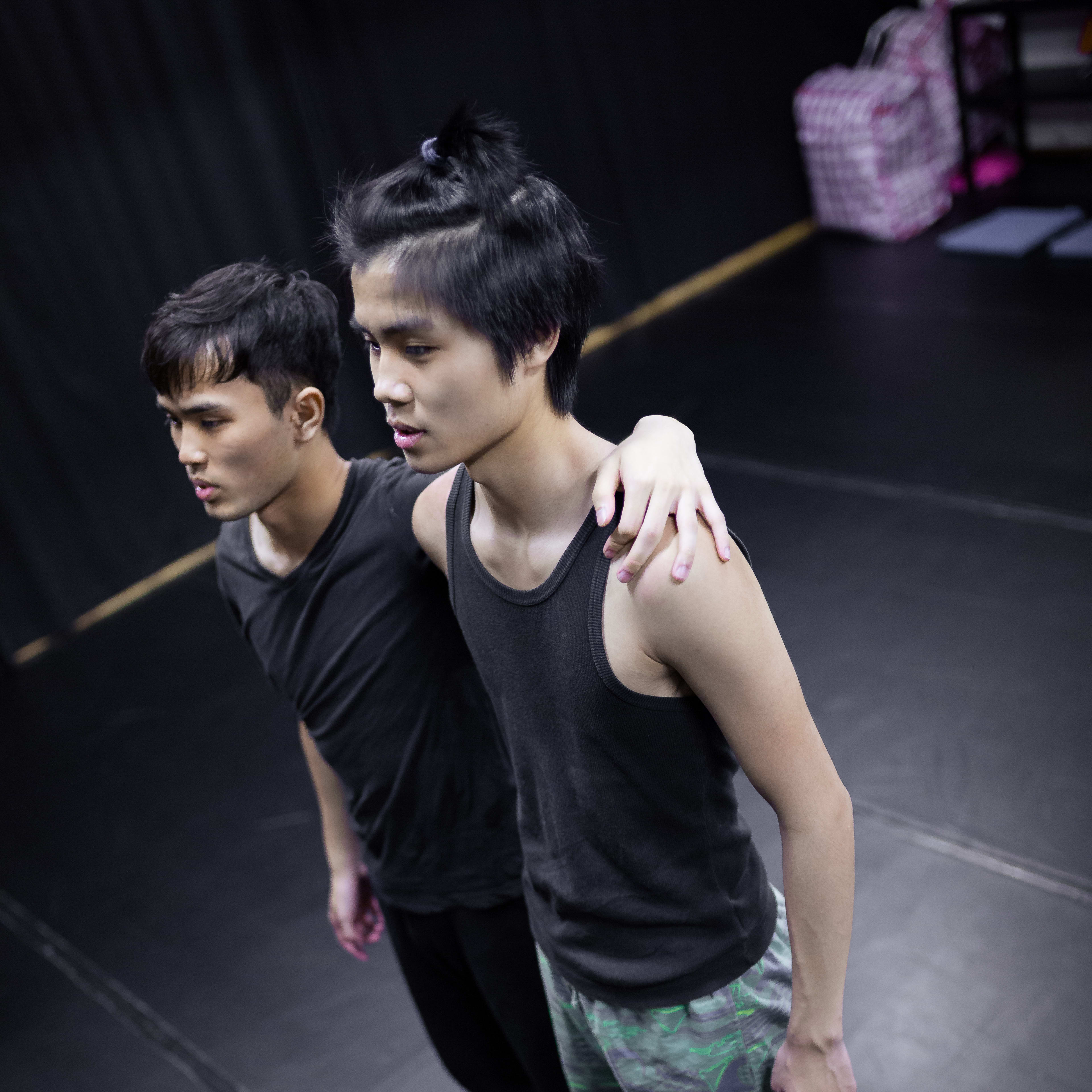 兩位男演員於「身心合一」（Psychophysical Actor Training）工作坊中進行肢體練習，專注於呼吸與重心運用。他們赤腳站在黑色舞台上，前方的演員身穿深色背心與花紋短褲，後方的夥伴則穿著黑色上衣與緊身褲，並將手搭在對方肩上作支撐。此排練場景展現了當代香港劇場對身心連結的嚴謹追求。「身心合一」工作坊於香港，2016年。