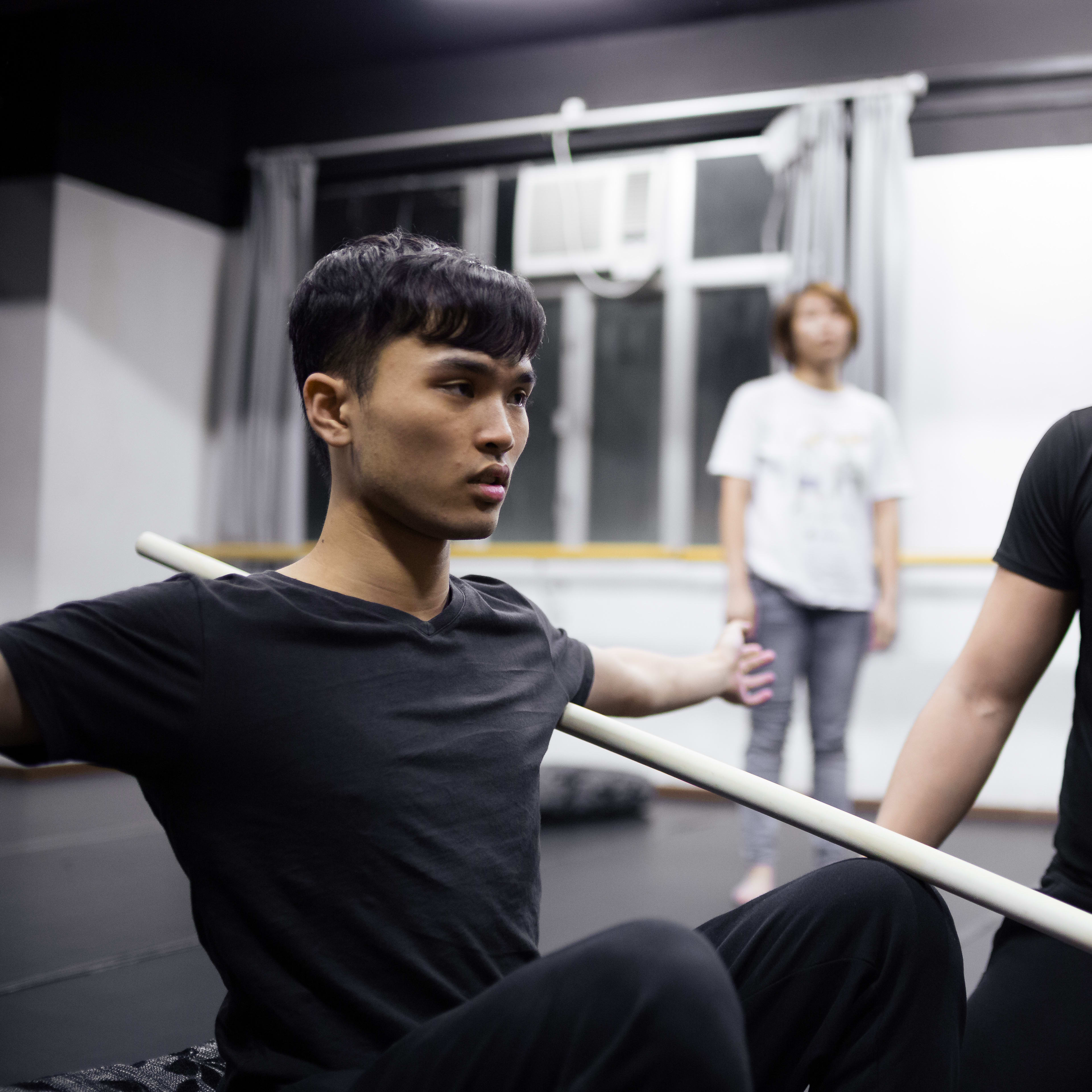 在排練室中，演員艾浩家（Ivor Houlker）正參與「身心合一」（Psychophysical Actor Training）香港演員工作坊。他盤坐在地，雙手橫握一根長棍置於肩後以調整姿態，旁邊的導師正專注地指導動作。這項專注於身體與意識連結的當代劇場訓練，體現了「身心合一」表演體系的核心。 「Psychophysical actor training workshops in Hong Kong, 2016」