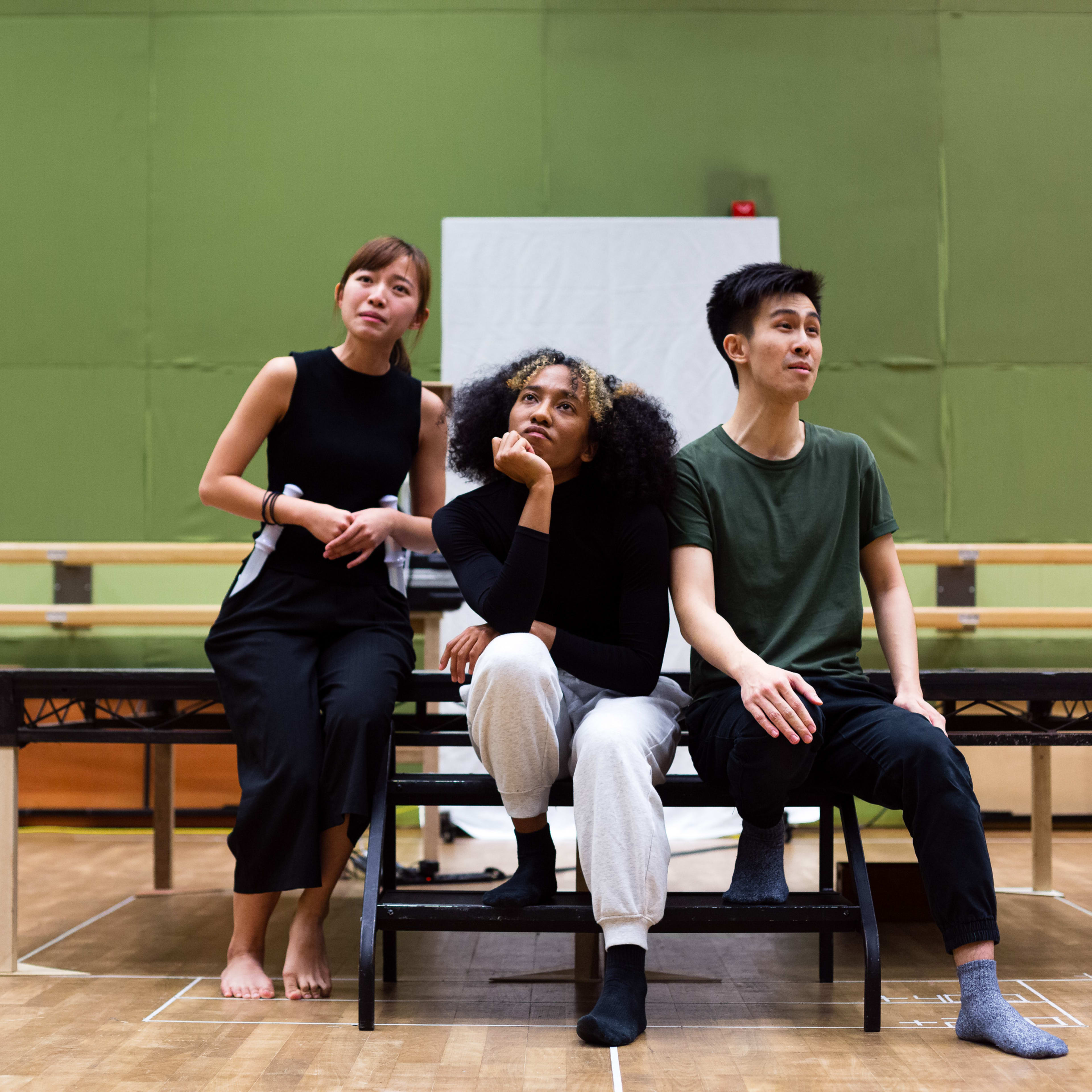 來自香港當代劇場團體「天台製作」(Rooftop Productions) 的三位演員，正為演出《天真與世故之歌》(Songs of Innocence and Experience) 進行排練。左側的冼樂欣（飾演夏娃／天使）仰頭沉思；中間的陳琬瑜（飾演撒旦／路西法）托腮思考；右側的張卓行（飾演天使）則坐姿端正，望向前方。場景設於簡約的排練室中，背後是綠色背景幕，展現了這部香港舞台製作的實驗性與排練現場的張力。《天真與世故之歌》排練