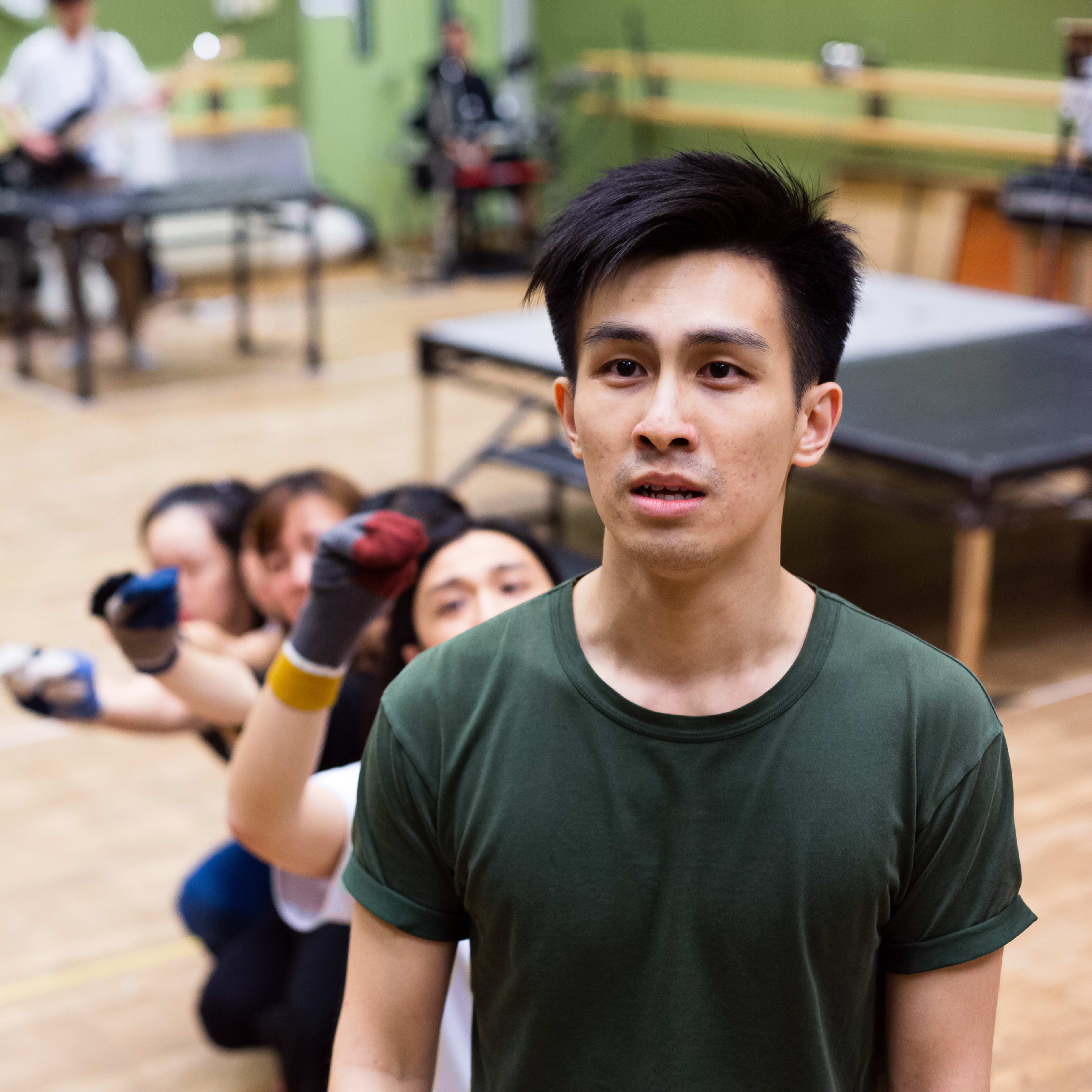在天台製作（Rooftop Productions）當代劇場作品《天真與世故之歌》的排練現場中，演員張卓行佇立於前景，表情專注而凝重。在他身後，包括麥晧鈿在內的一眾演員排成一列，透過層層疊疊的手部動作，呈現出充滿意象的肢體編排或集體定格。排練室背景僅有簡約木凳與樂器，真實呈現香港劇場排練時那份原始且實驗性的張力。《天真與世故之歌》排練。