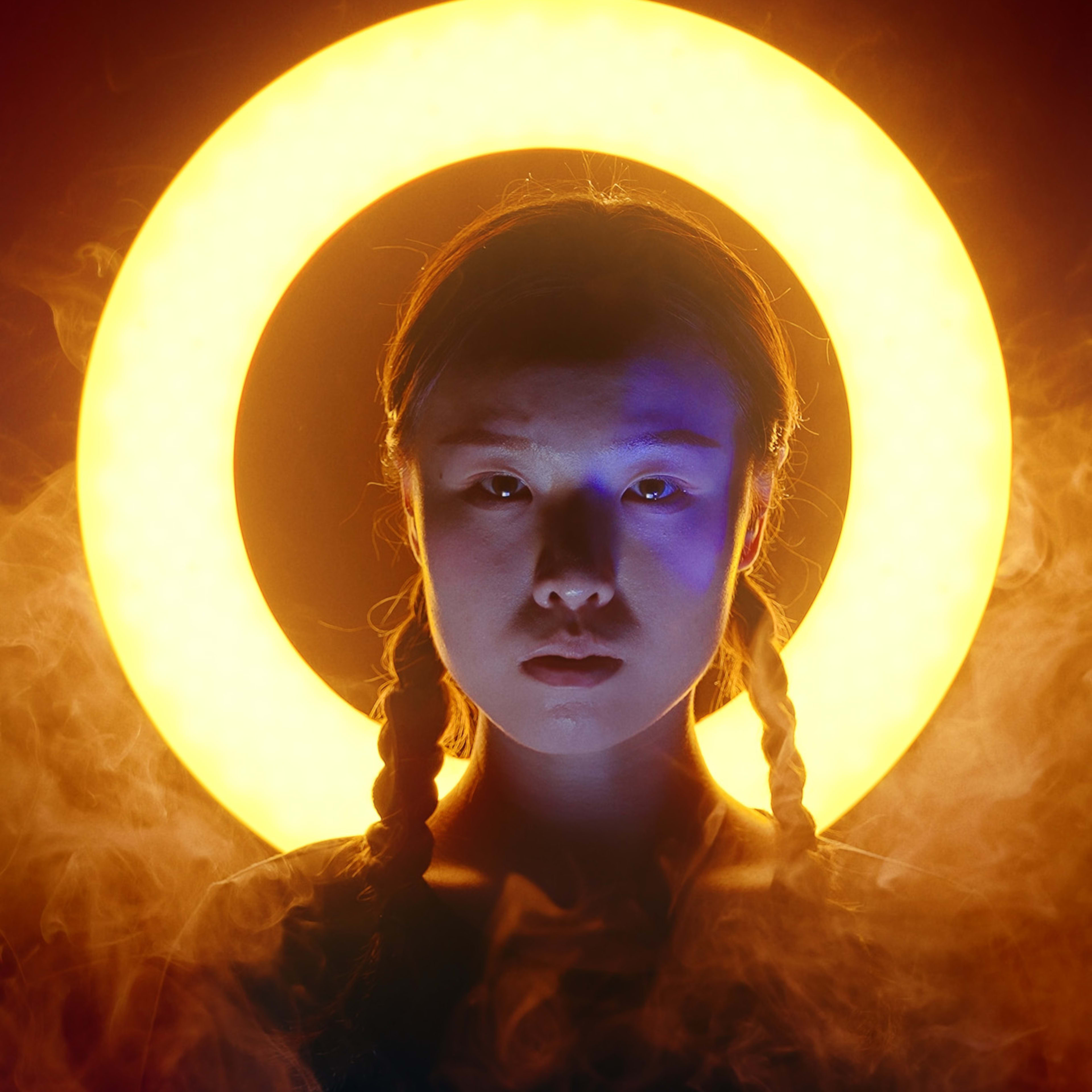 天台製作（Rooftop Productions）當代劇場作品《天真與世故之歌》的劇照，演員沈卓瑤（飾演阿必迭）正襟危坐，神情肅穆。她身後環繞著一圈如光環般的金色光影，周圍瀰漫著繚繞煙霧，光影對比強烈，展現出舞台表演的戲劇張力與深沉情緒。《天真與世故之歌》西九文化區