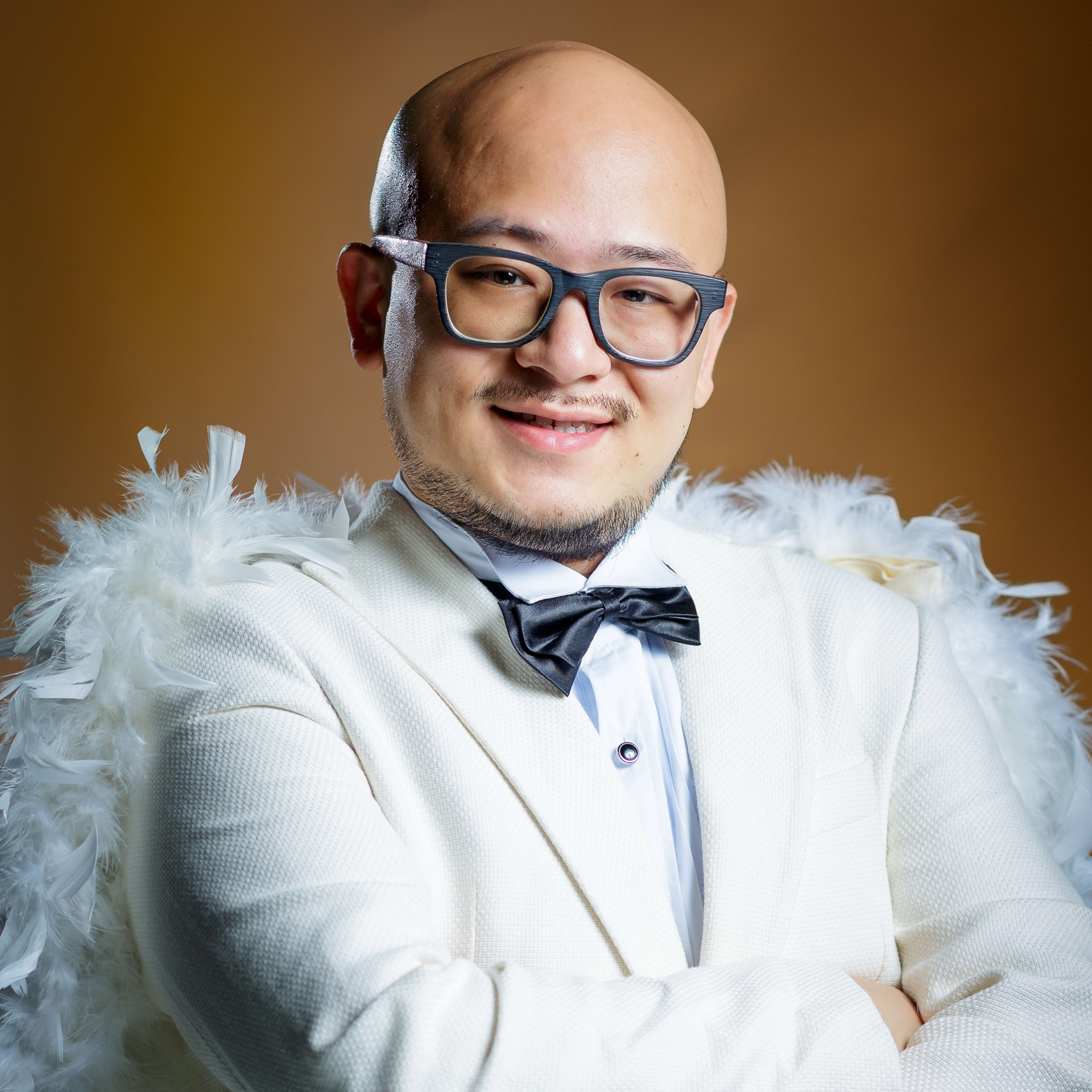 Portrait of performer Alfie Leung for the Rooftop Productions contemporary Hong Kong theatre show 'Superheroes Don't Give a Shit'. Leung is wearing a white tuxedo jacket paired with a white feather boa, styled as a whimsical, satirical take on a superhero costume. He is captured in a studio setting with warm professional lighting, posing with his arms crossed and a gentle, inviting smile. 'Superheroes Don't Give a Shit!'