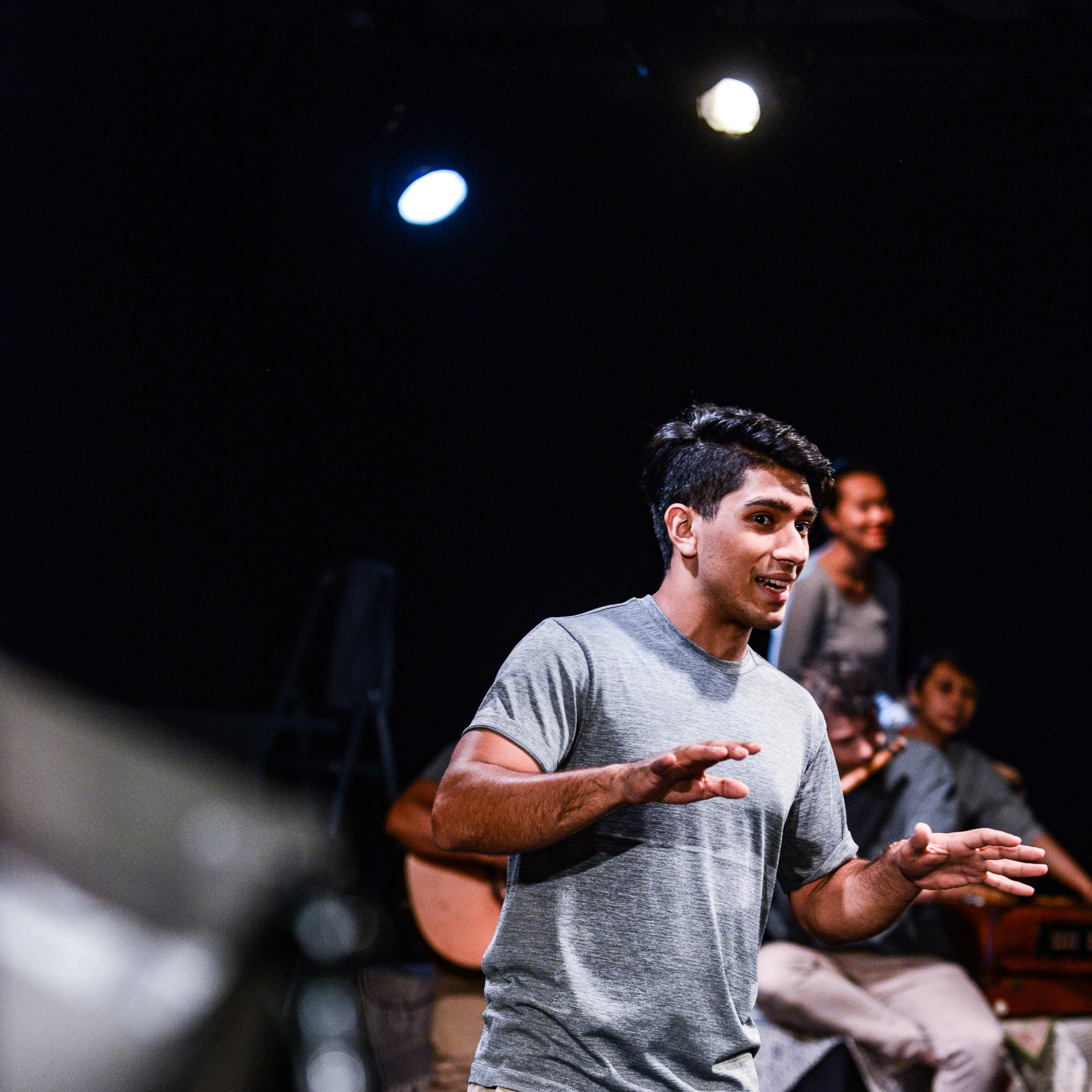 在天台製作 (Rooftop Productions) 當代劇場作品《山下的證詞》中，演員巫加沙 (Mohammad Kashif Ali) 站在聚光燈下，身穿灰色T恤與淺色長褲，正投入地表演並以手勢輔助敘事。背景可見其他演員與樂器，營造出充滿張力的舞台氛圍。《山下的證詞》