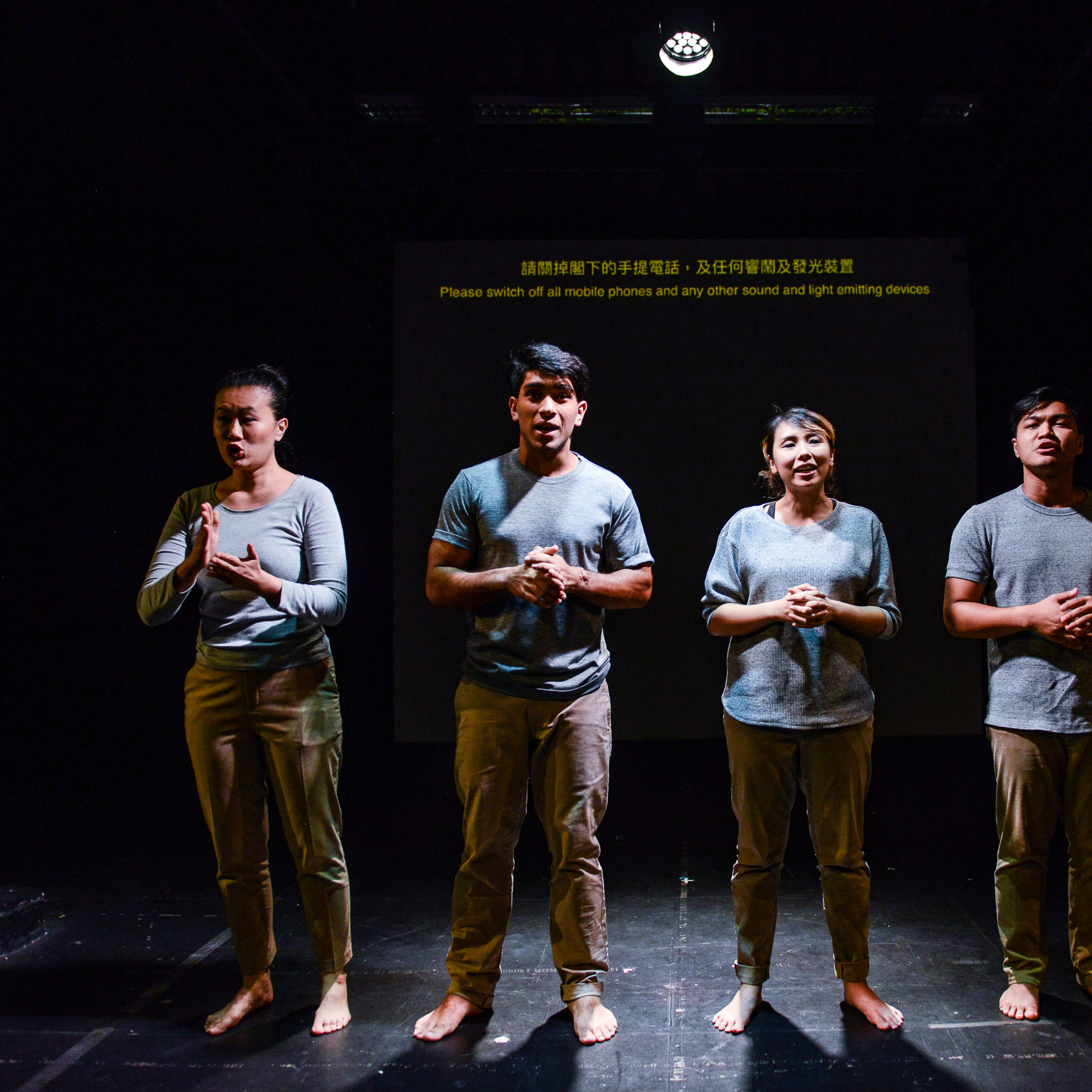 四位演員在香港當代劇場《山下的證詞》排練現場，並排站立於舞台中央。他們身穿休閒大地色系服裝，在頂部聚光燈的照射下，展現出極簡而充滿張力的戲劇氛圍。演員們（左起：李婉晶、Basnet Tusar、李里莎、巫加沙）表情嚴肅凝重，深刻詮釋出劇中沉重而發人深省的主題。他們身後的螢幕模糊地顯示著中英文提示，提醒觀眾關閉電子設備。《山下的證詞》