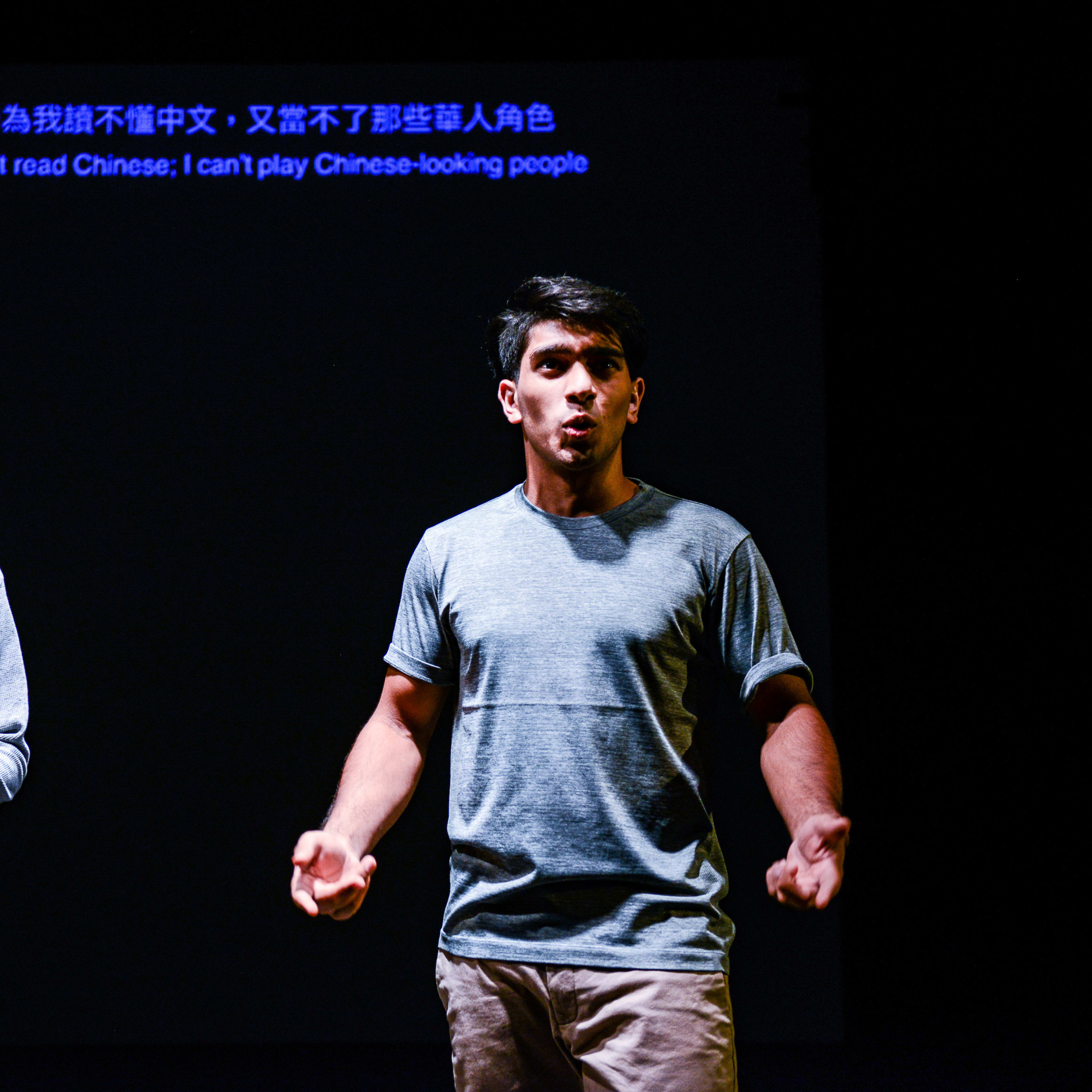 在天台製作（Rooftop Productions）的當代劇場作品《山下的證詞》演出中，演員巫加沙（Mohammad Kashif Ali）與吳景隆站在昏暗的舞台上，背景為純黑色。舞台上方投影出字幕：「因為我讀不懂中文，又當不了那些華人角色」，此幕展現了該劇探討文化認同與身份定位的深刻主題。