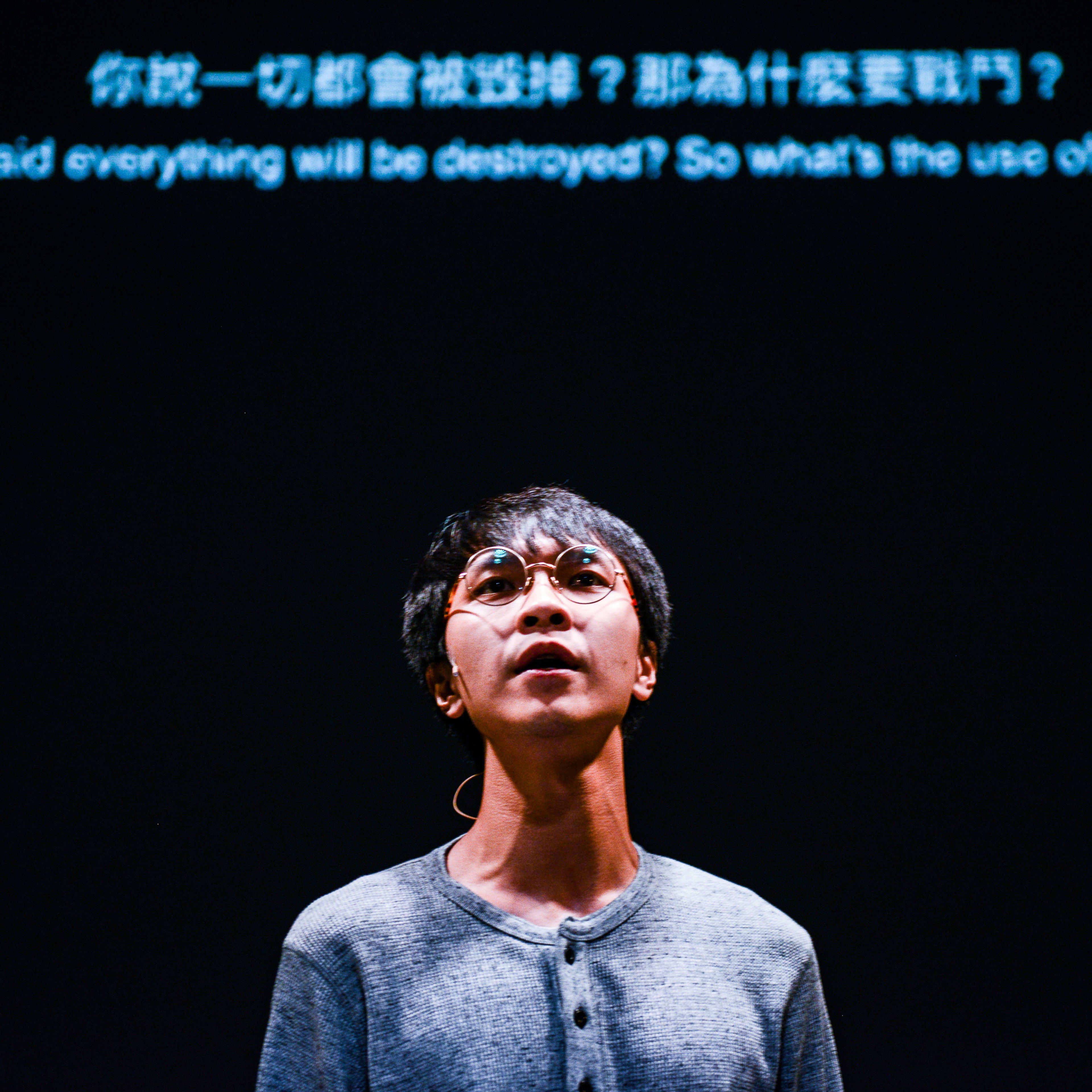 天台製作（Rooftop Productions）當代劇場作品《山下的證詞》（Testimony）的戲劇性舞台攝影。演員吳景隆在黑暗的背景中被單一聚光燈照亮，他佩戴圓框眼鏡，身穿休閒灰衫，神情深沉地仰望。在他身後的投影字幕寫道：「你說一切都會被毀掉？那為什麼要戰鬥？」